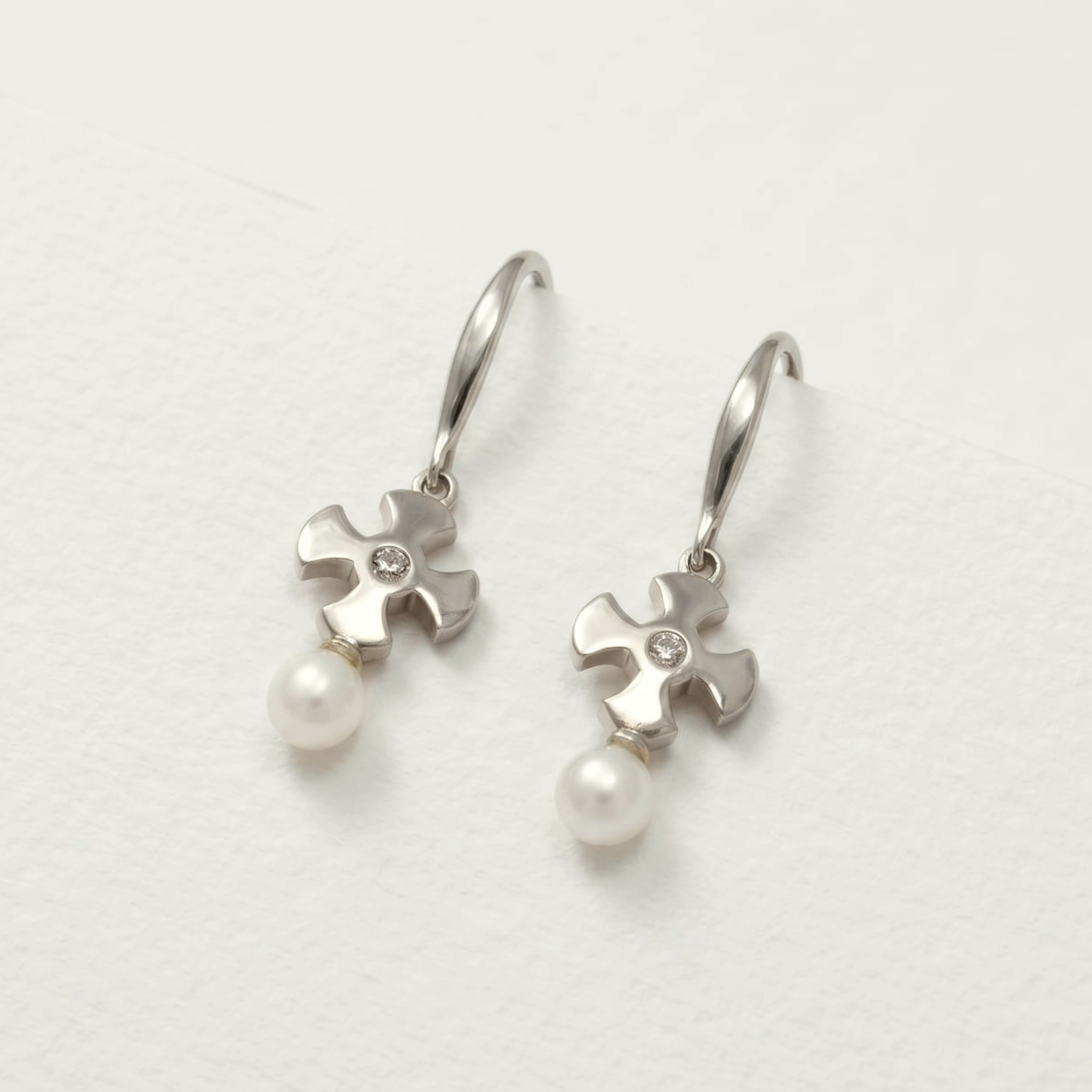 Pair of sterling silver drop earrings featuring sculptural St Cuthbert crosses accented with freshwater pearls and a small clear stone, styled on a soft beige background.