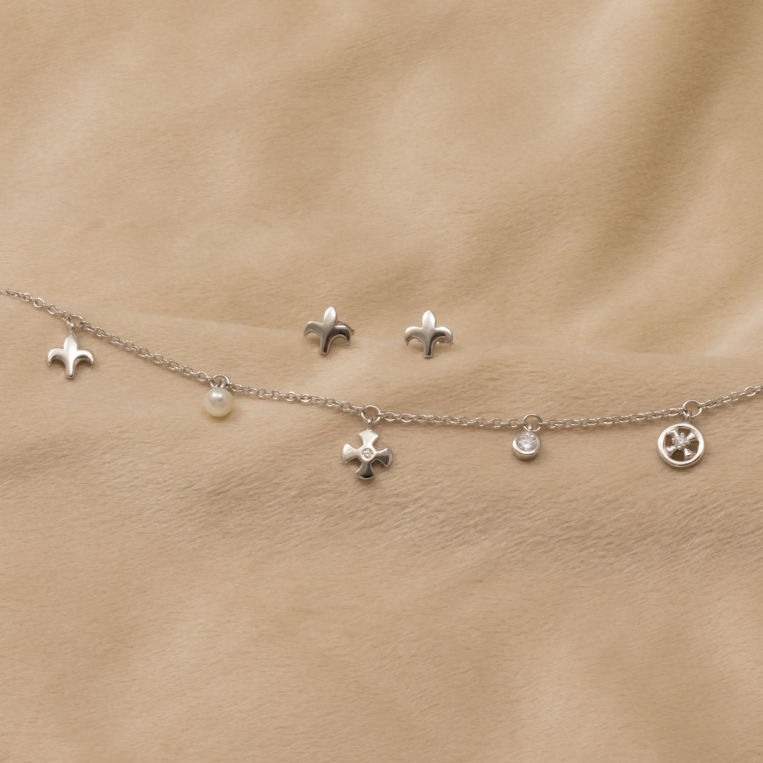 Delicate sterling silver charm bracelet featuring a fleur de lis, cross, pearl, and sparkling stone accents, accompanied by matching fleur de lis stud earrings.