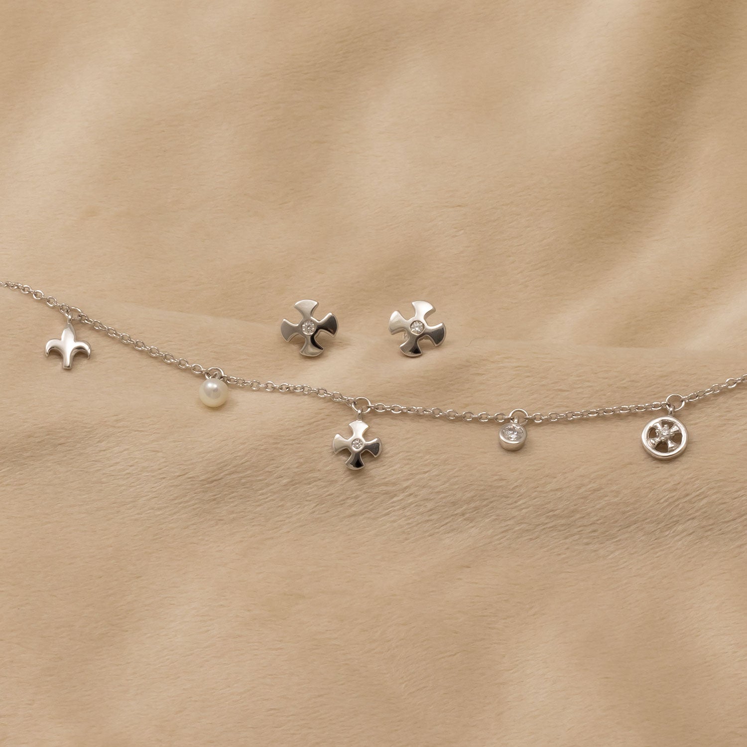 Pair of silver stud earrings designed with a sculptural fleur de lis motif, styled alongside a delicate sterling silver charm bracelet featuring mixed pendants and a small pearl.