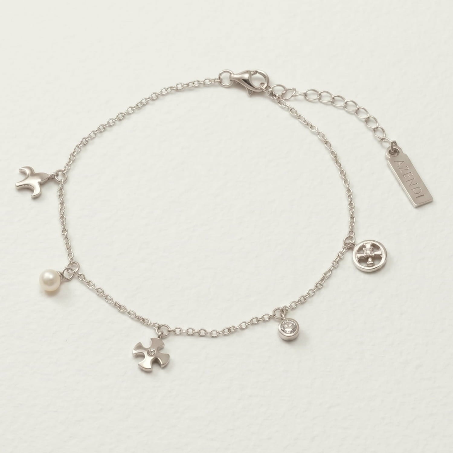 Delicate sterling silver bracelet featuring a collection of small charms including a cross, fleur de lis, pearl, and crystal accents on an adjustable chain with a branded tag.