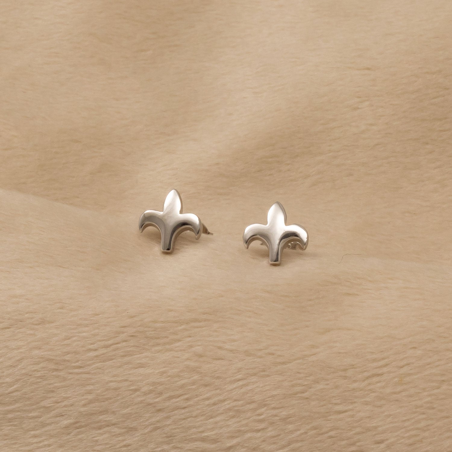 Pair of sterling silver stud earrings shaped as smooth Fleur de Lis motifs, displayed on a soft beige fabric background emphasizing their polished finish.