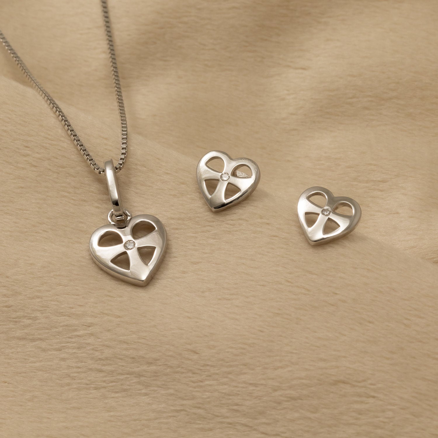 Sterling silver heart-shaped pendant and matching stud earrings featuring a sculptural cross design, displayed on a soft beige fabric background.
