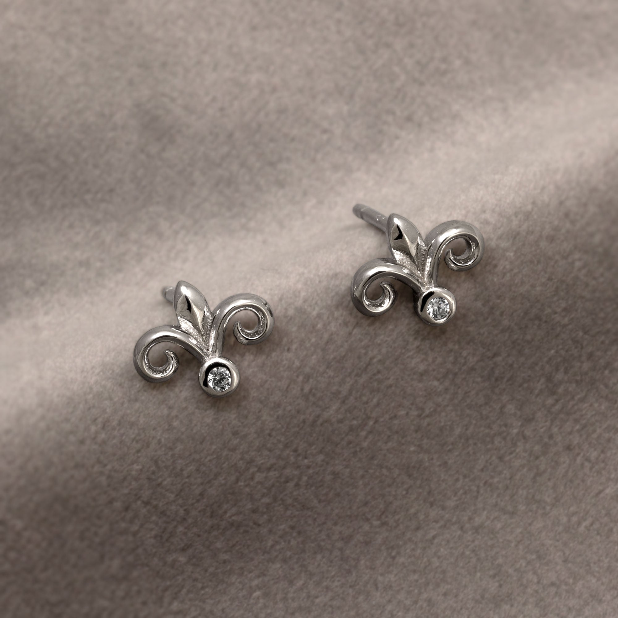 Pair of sterling silver stud earrings shaped as fleur de lis motifs, each featuring a polished finish and a small clear stone set at the base, displayed on a white background. Place on a grey velvet cloth.