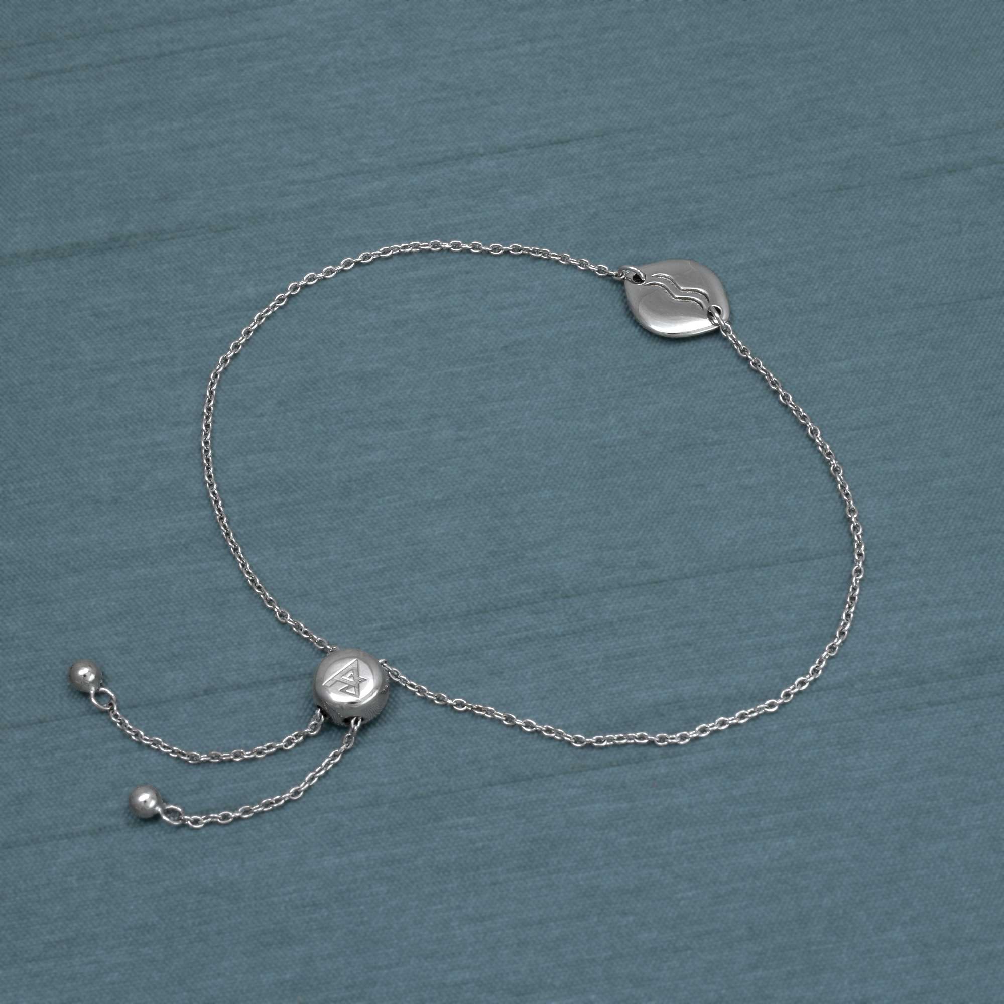 Delicate sterling silver adjustable bracelet featuring a sculptural Aquarius zodiac charm and sliding bead closure, styled on a muted blue fabric background.
