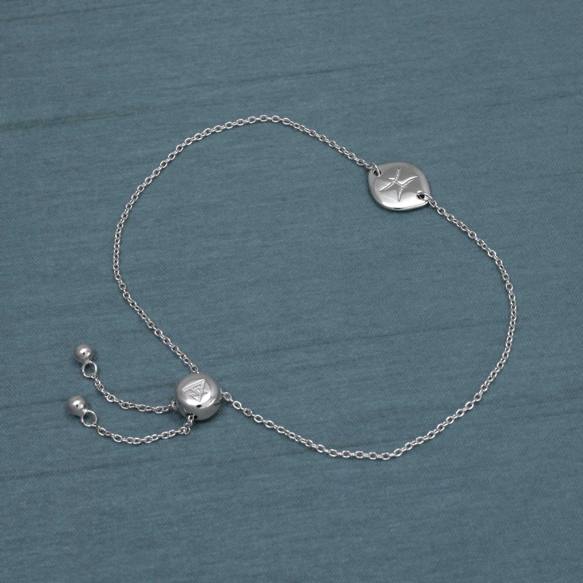 Delicate sterling silver adjustable bracelet with a circular Pisces zodiac charm, featuring a sliding bead for customizable fit, displayed on a muted blue fabric background.