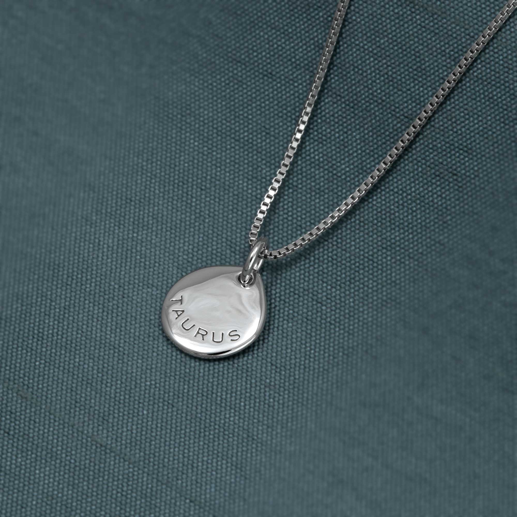 Delicate sterling silver pendant engraved with the word Taurus, hanging from a fine box chain against a textured teal fabric background.