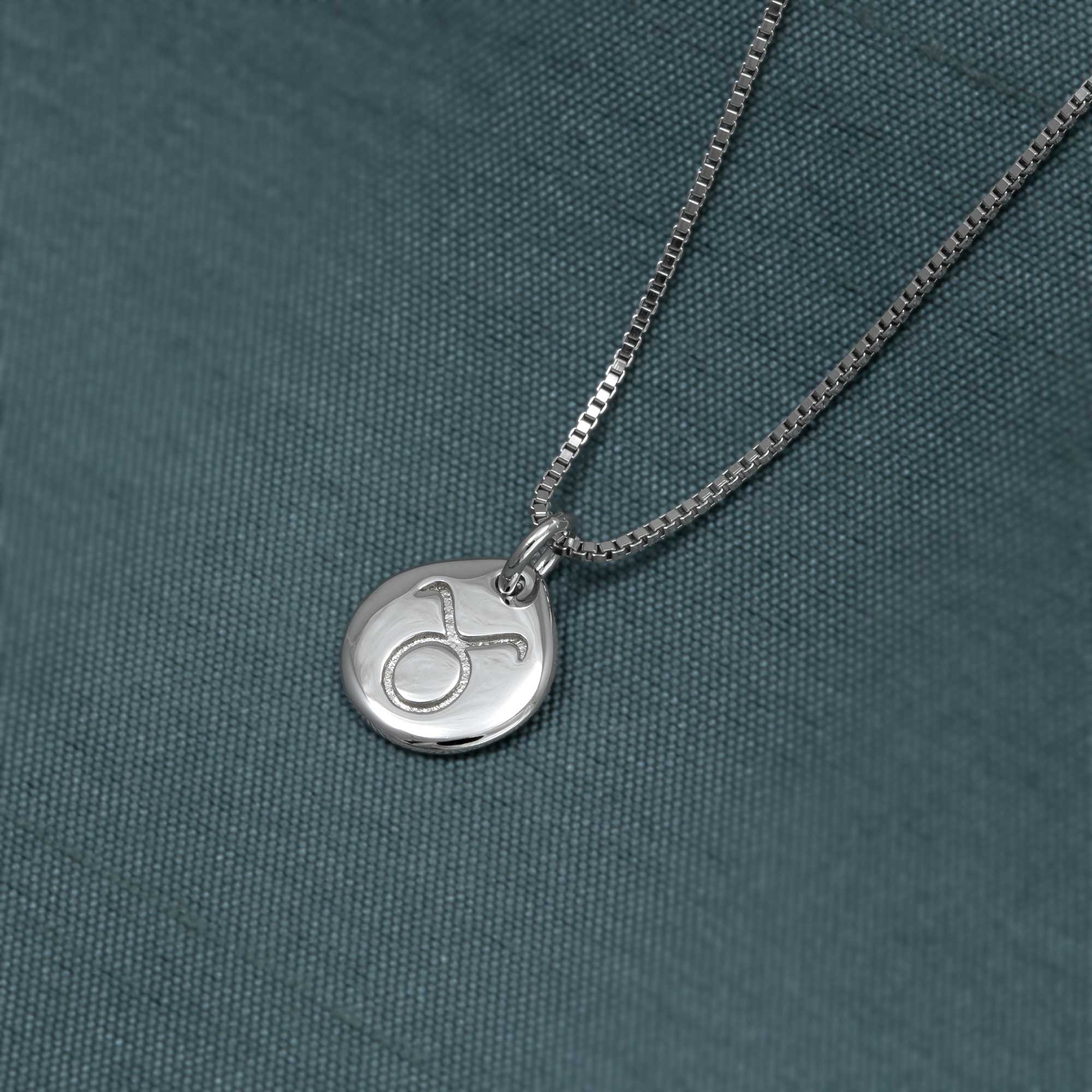 Delicate sterling silver pendant featuring a sculptural Taurus zodiac symbol, suspended from a refined box chain, displayed on a textured teal fabric background.