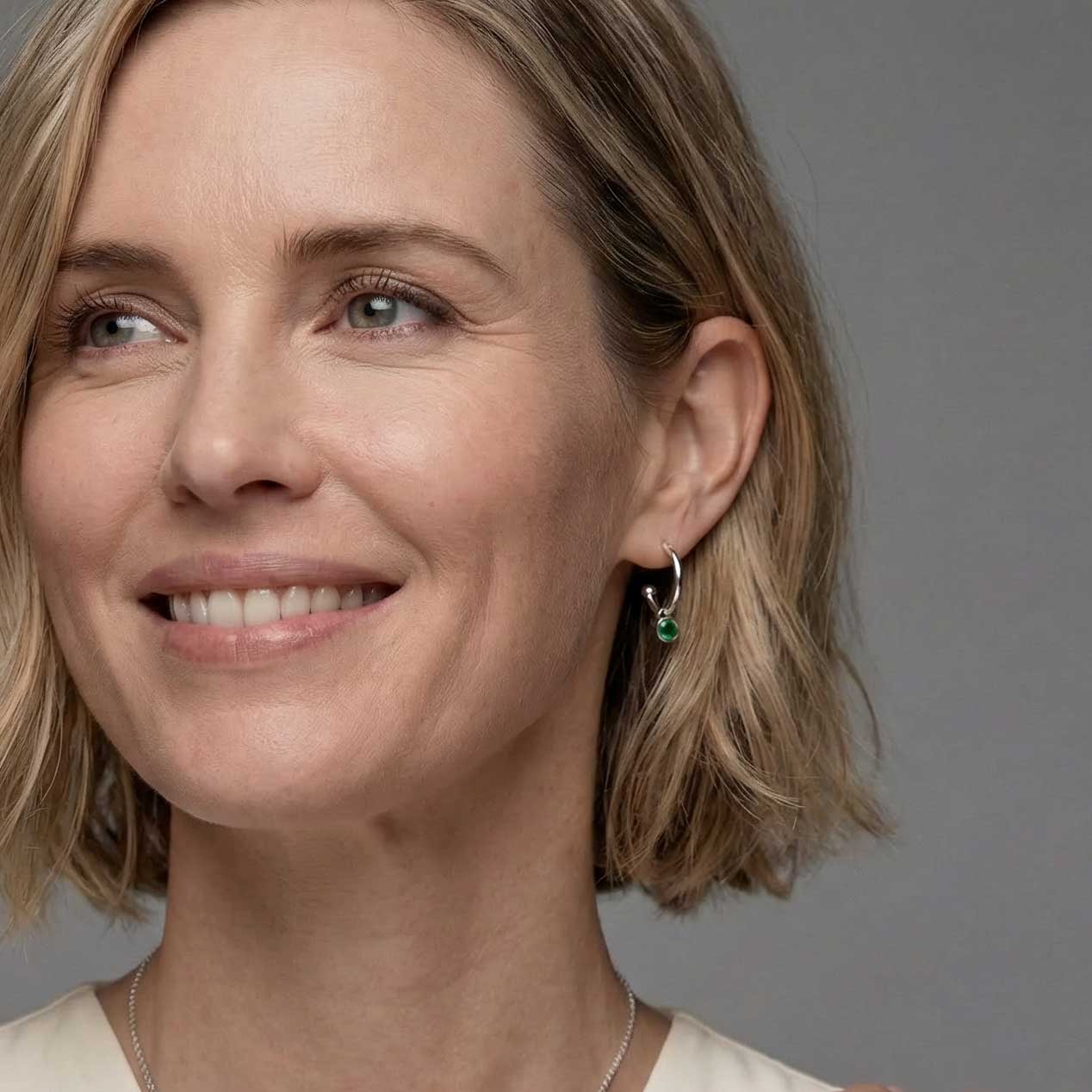 Delicate sterling silver hoop earrings featuring a vibrant Siberian emerald birthstone, highlighted by a sleek, contemporary design worn by a smiling woman.