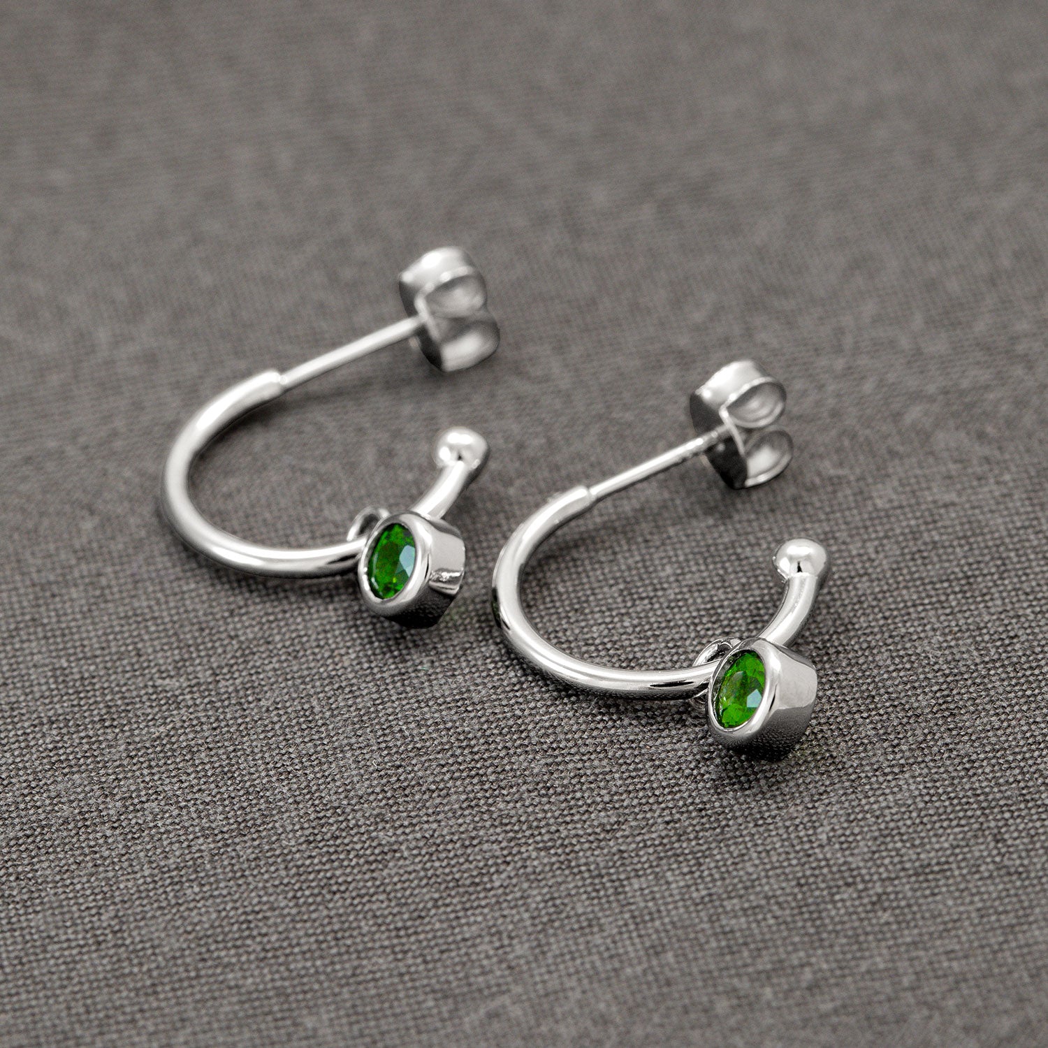 Delicate sterling silver hoop earrings featuring bezel-set Siberian emerald gemstones, designed with a modern sculptural curve and secured with butterfly backs on a textured grey fabric.