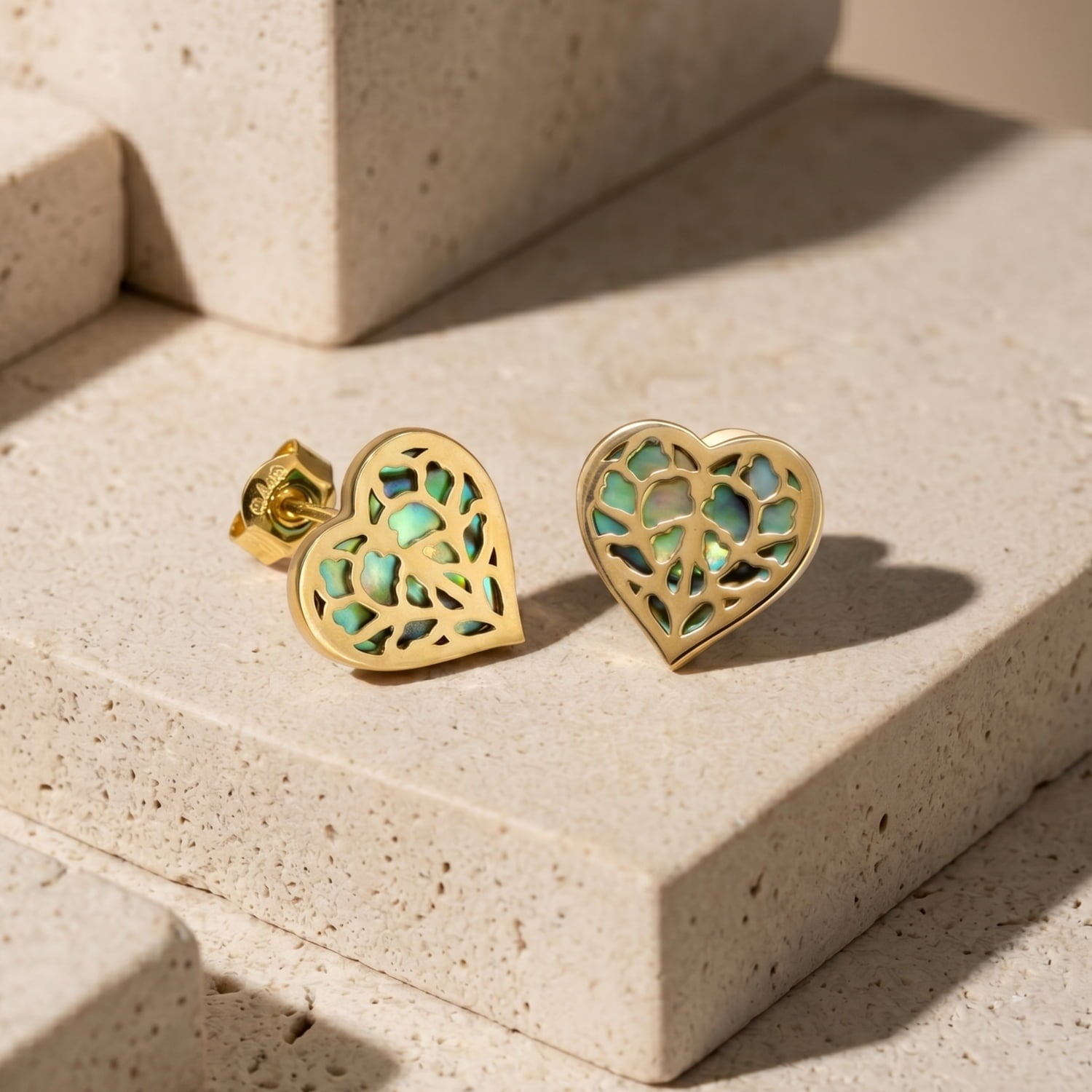 9 carat gold stud earrings featuring a heart-shaped openwork design with iridescent abalone inlay, laid on a polished stone background