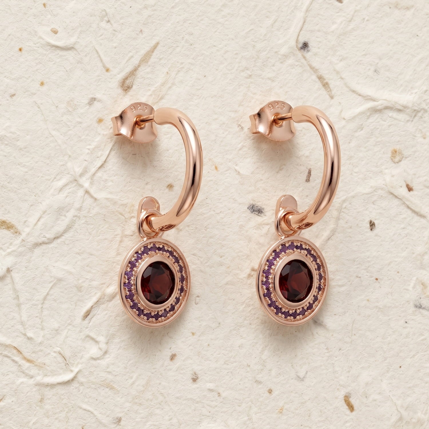 Pair of rose gold vermeil hoop earrings featuring oval garnet and amethyst gemstones set within decorative bezels, showcasing an elegant and sculptural design.