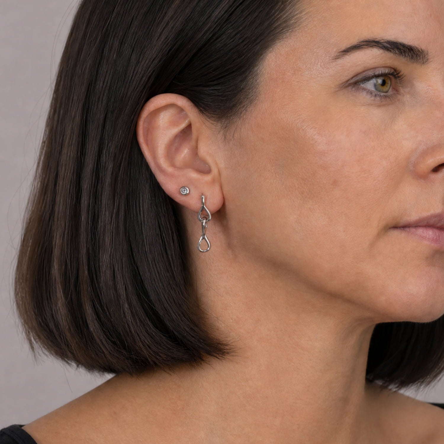 A close-up side profile of a woman with dark brown hair wearing a delicate silver drop linked earrings and a small silver stud earring against a neutral light gray background.