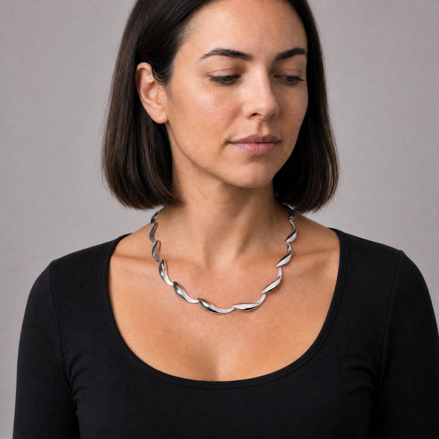 A woman with short dark brown hair wearing a black scoop-neck top and a twisted silver chain necklace, photographed against a neutral gray background.