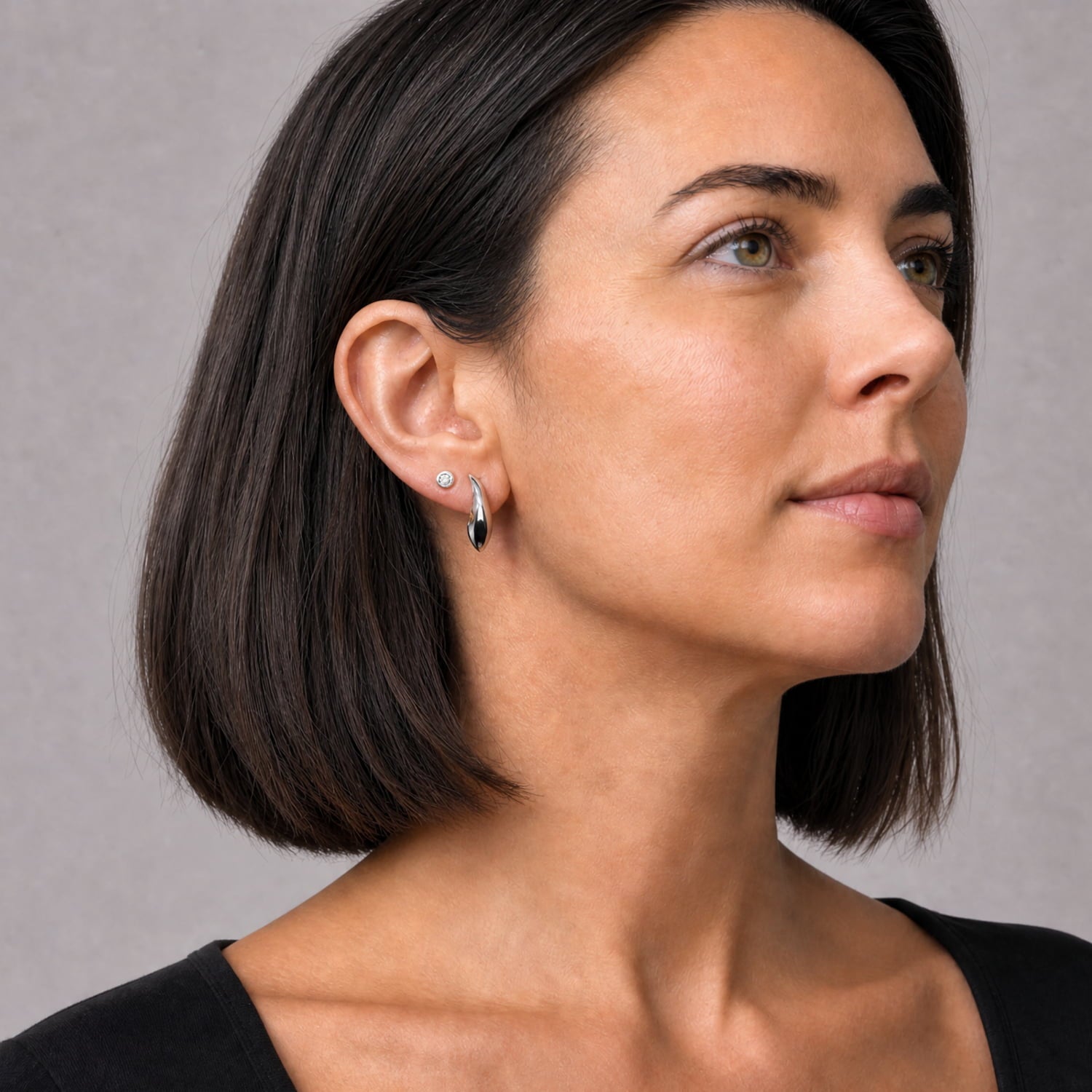 A close-up side profile of a woman with a short dark bob, wearing silver curve stud earring with a black stone on the lobe and a small round stud earring, against a neutral gray background.