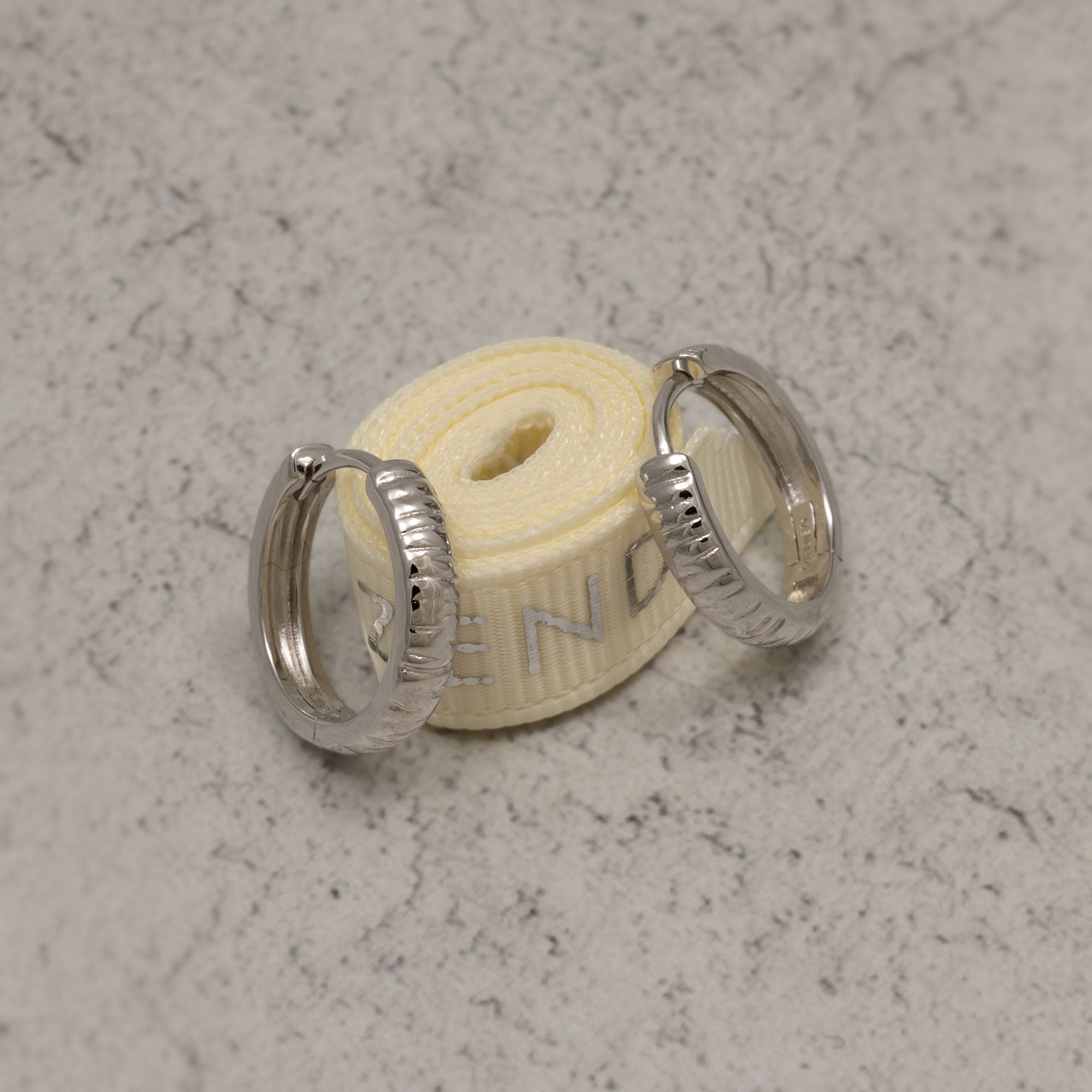 Pair of sterling silver hoop earrings featuring a grooved textured design with hinge closures, showcasing a contemporary and sculptural style on a white background. Placed on a marble base.