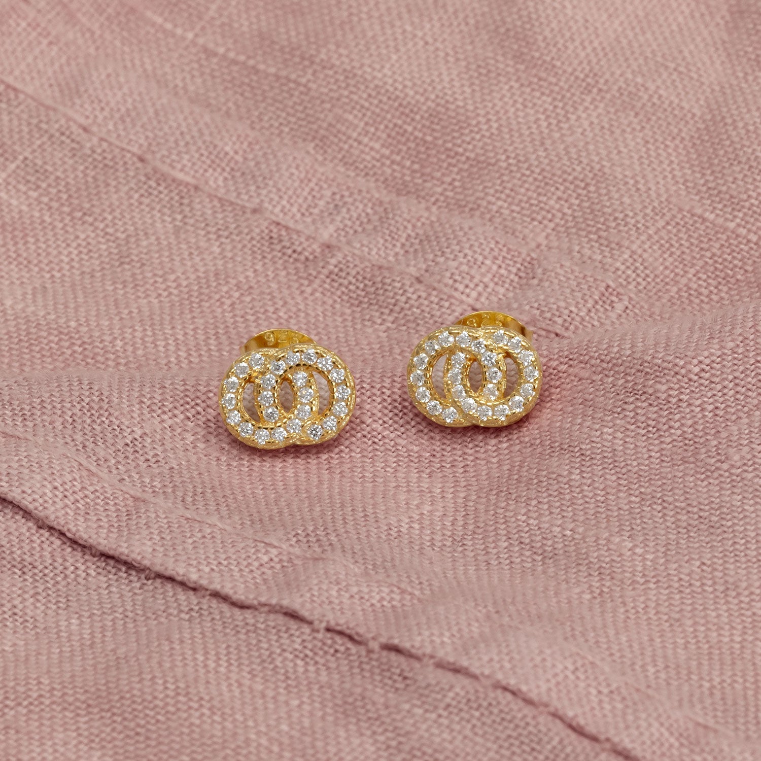 Pair of yellow gold vermeil stud earrings featuring interlocking circles paved with sparkling cubic zirconia, displayed on a soft textured fabric background.