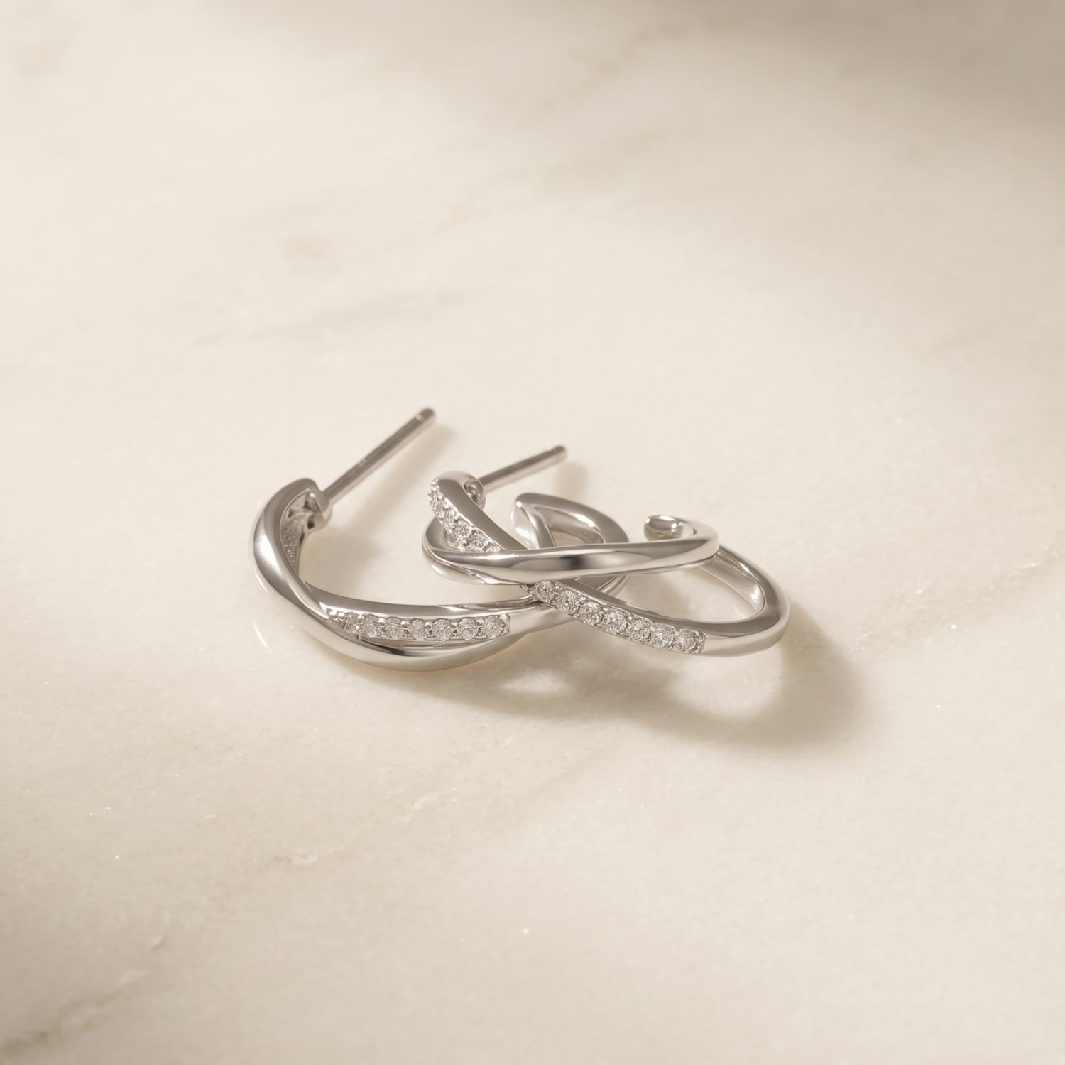 Pair of sterling silver hoop earrings featuring an elegant crossover wave design accented with pavé-set stones, laid upon a marble background.