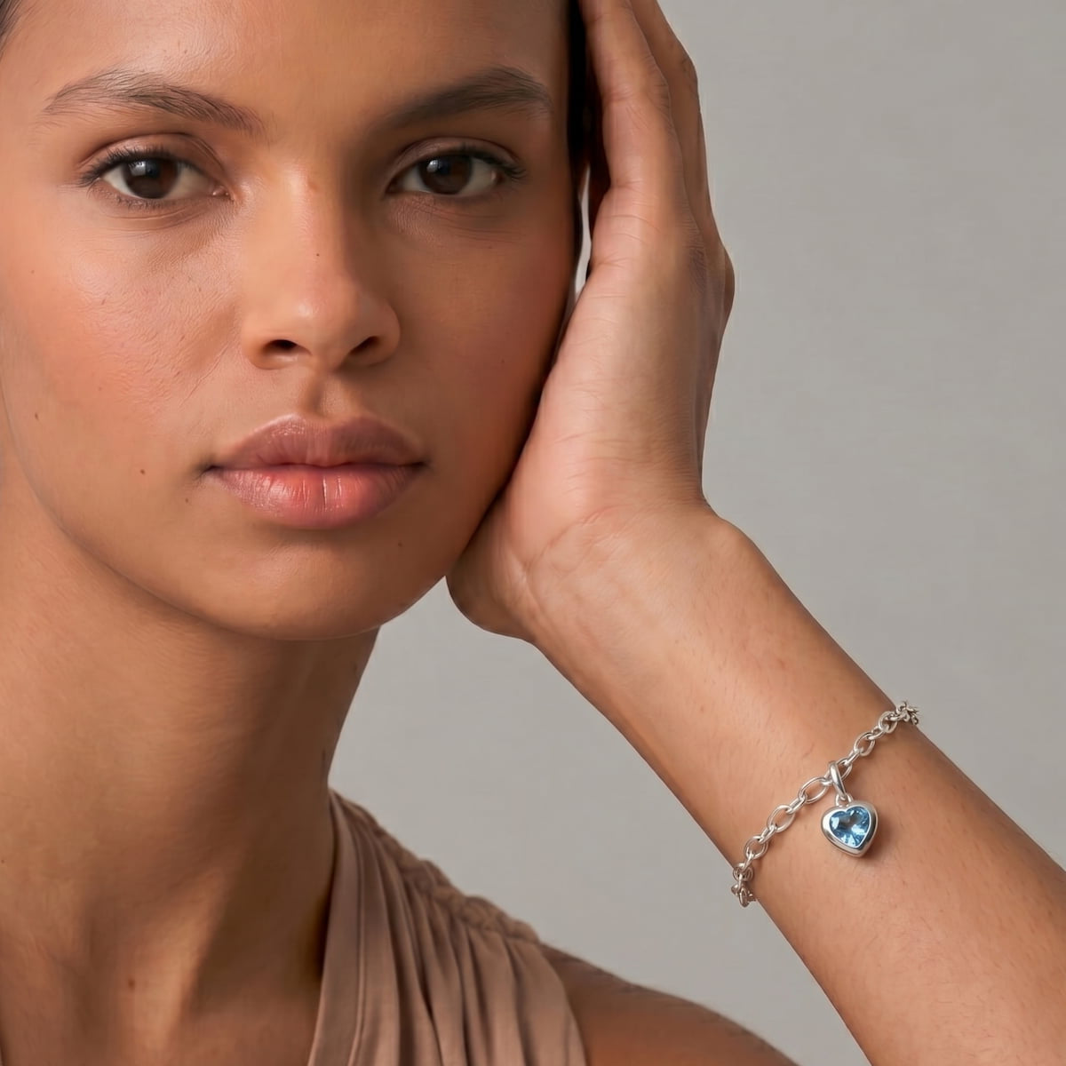 Close-up of a woman wearing a sterling silver chain bracelet featuring a blue topaz heart charm, styled with a neutral-toned sleeveless top against a soft grey background.