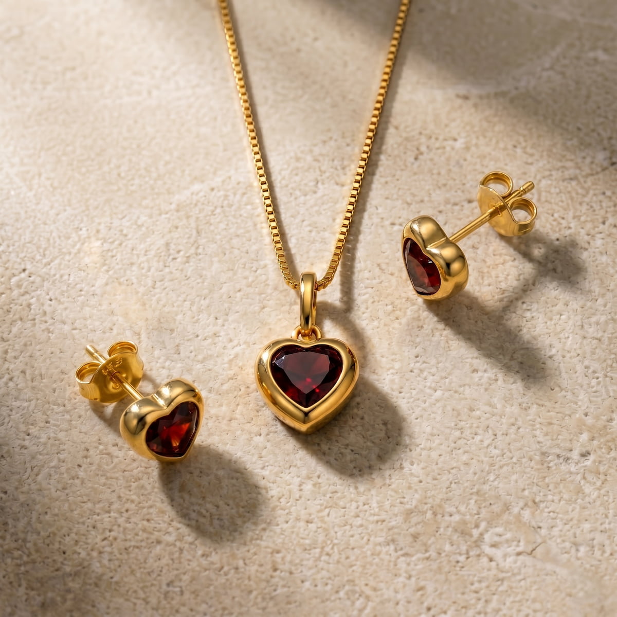 Gold vermeil jewellery set with heart-shaped garnet pendant and earrings on a textured beige surface