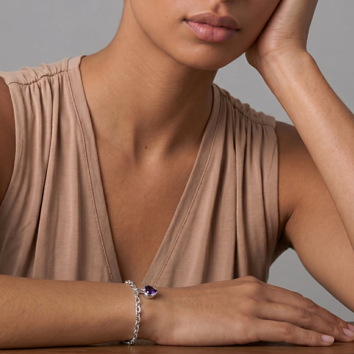 Close-up of a woman wearing a sterling silver chain bracelet featuring a sculptural amethyst heart charm, styled with a neutral-toned sleeveless top against a soft grey backdrop.