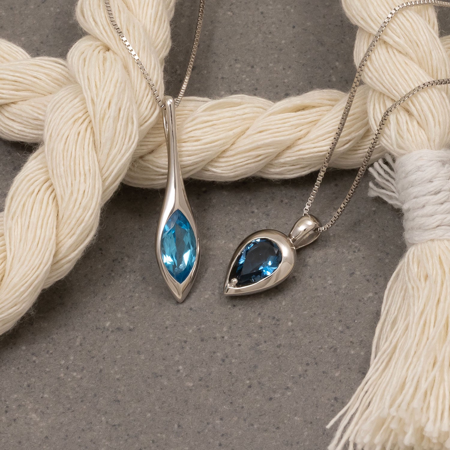 Two silver pendants with blue gemstones on a textured surface with white yarn.
