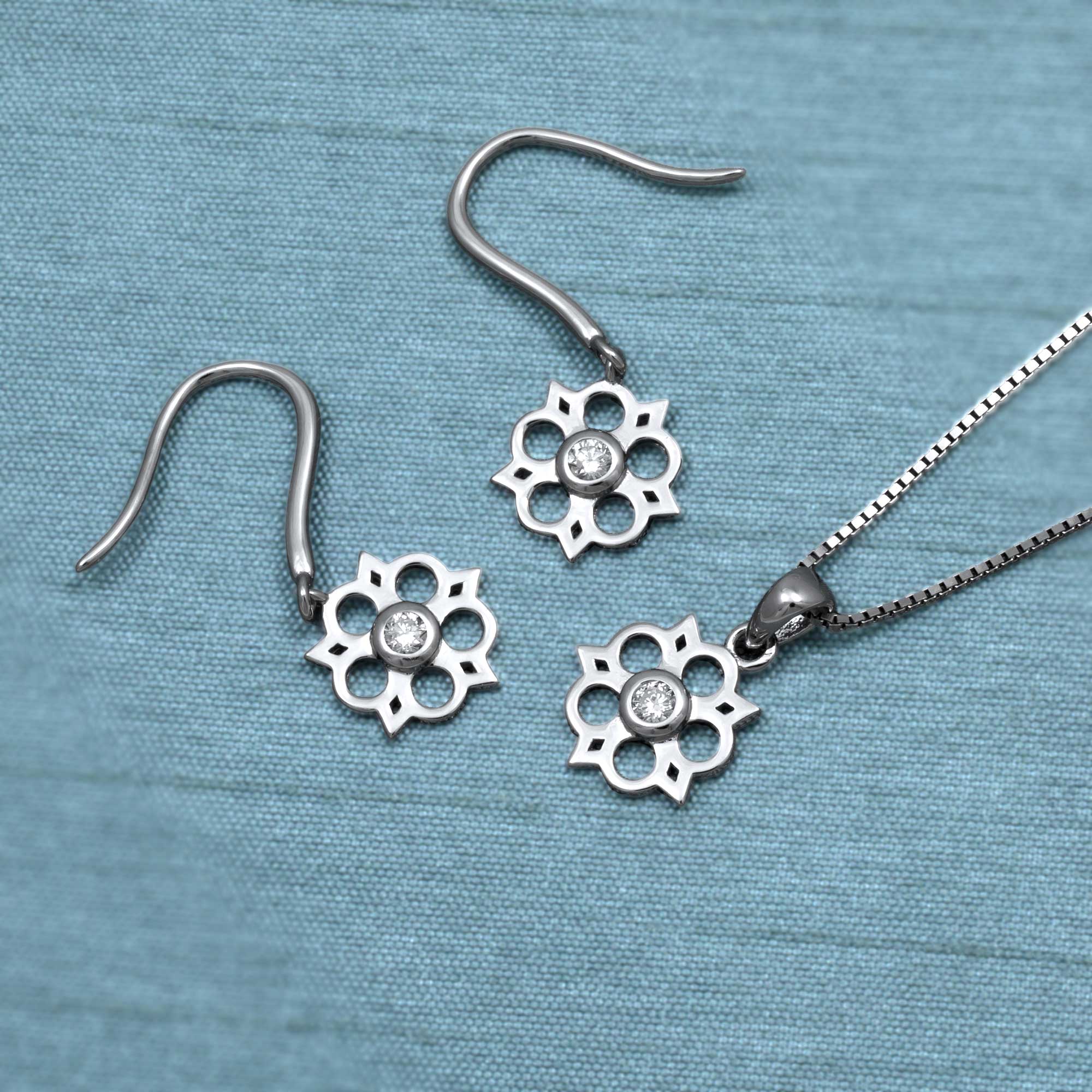 Sterling silver Yorkshire rose pendant featuring a 0.07 carat lab-grown diamond at the centre, suspended on a delicate fine chain with sculptural elegance. Delicate sterling silver drop earrings featuring an openwork Yorkshire rose motif with a central lab-grown diamond. Place on a teal satin cloth.