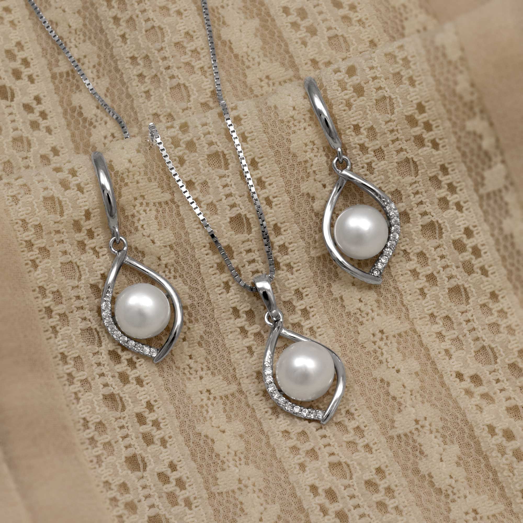 Sterling silver drop earrings and matching pendant featuring open twisting frames set with pavé cubic zirconia and a central freshwater pearl, styled on textured fabric.