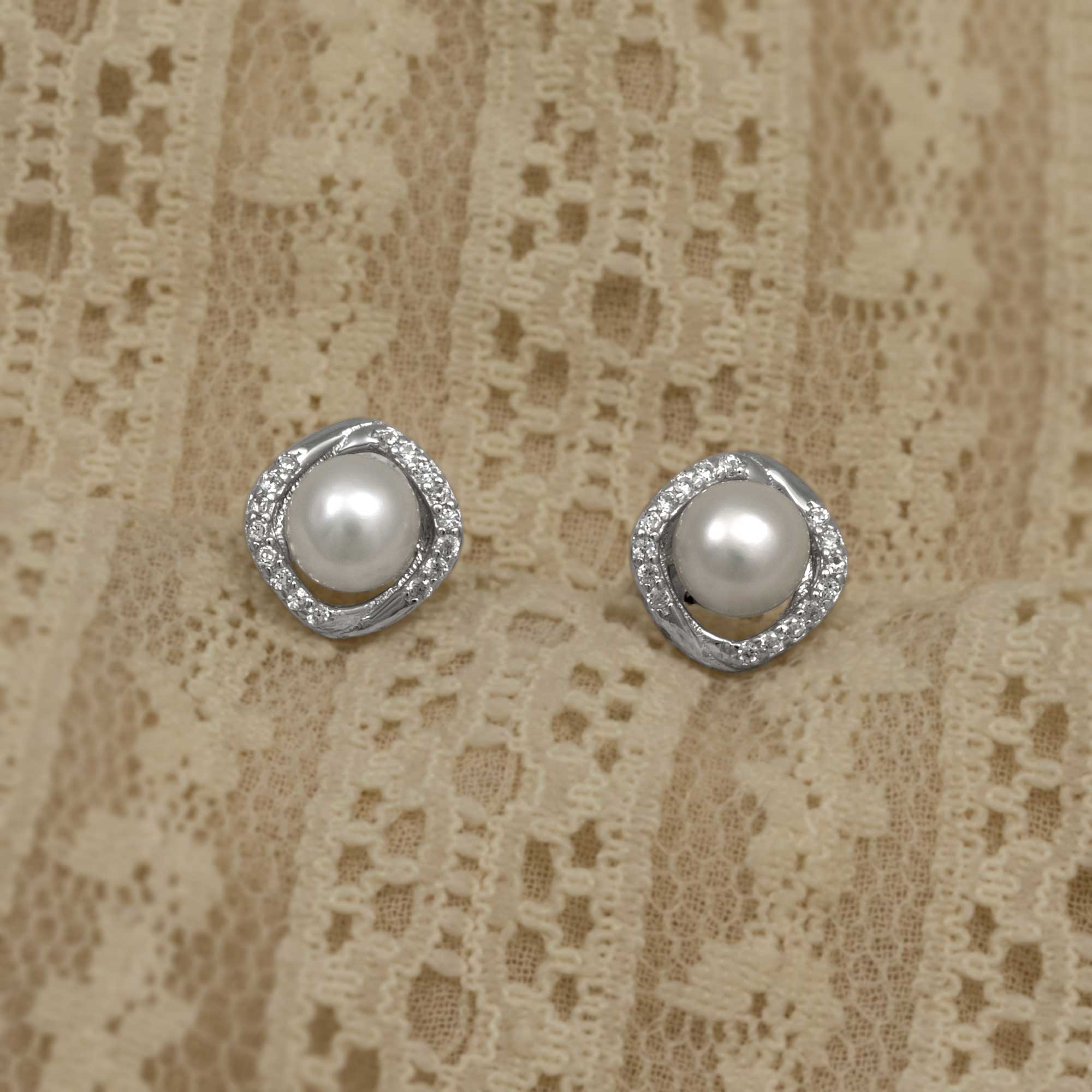 Pair of sterling silver stud earrings featuring round freshwater pearls encircled by pavé-set cubic zirconia in an elegant wraparound design on a soft textured fabric background.