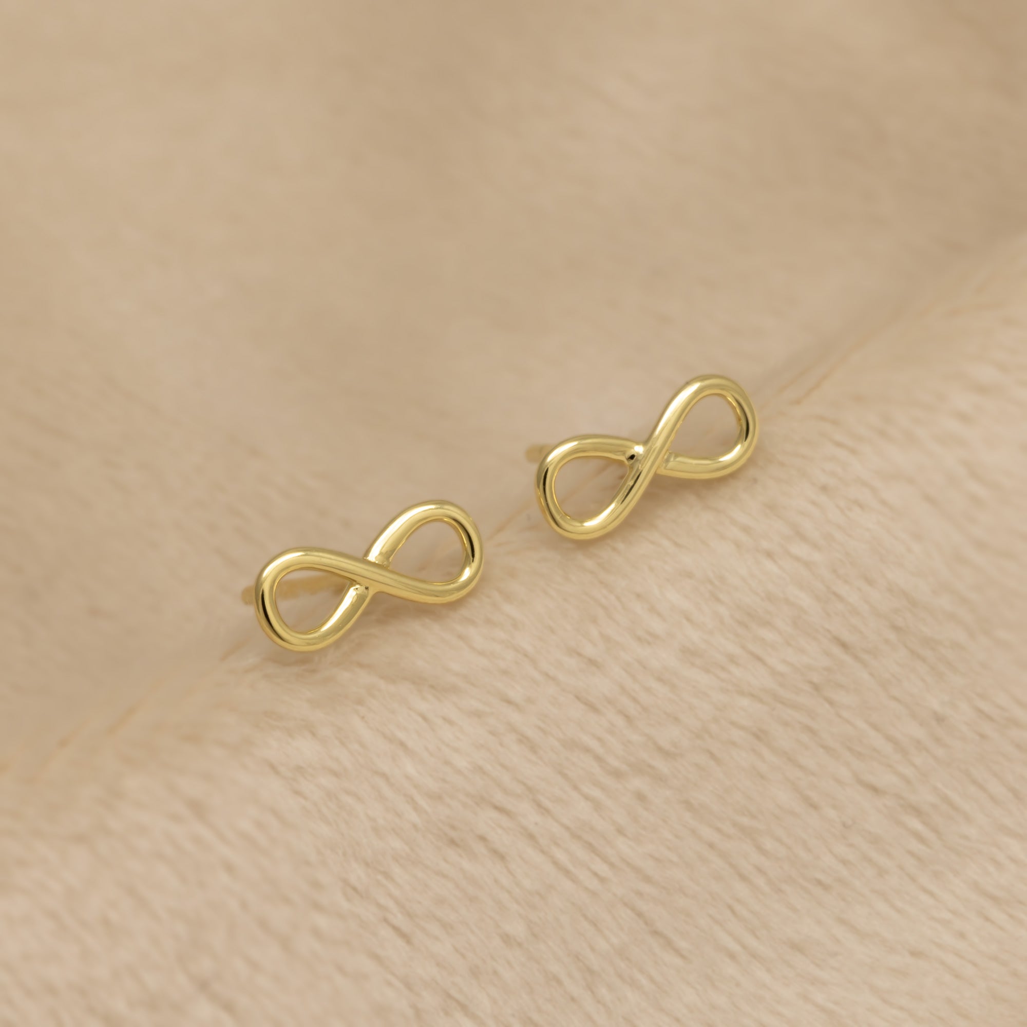 Gold vermeil infinity symbol stud earrings on a beige background