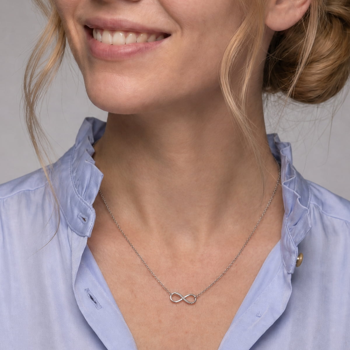 A close-up of a woman wearing a delicate silver infinity necklace with lab grown diamonds, styled with a light blue button-down shirt.