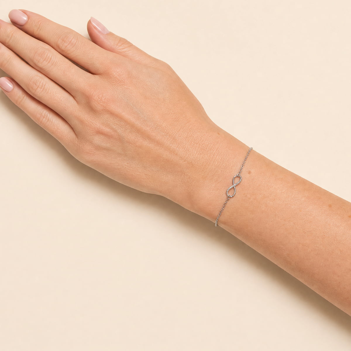 A delicate silver infinity bracelet, set with lab grown diamonds, worn on a person's wrist, shown against a soft beige background.