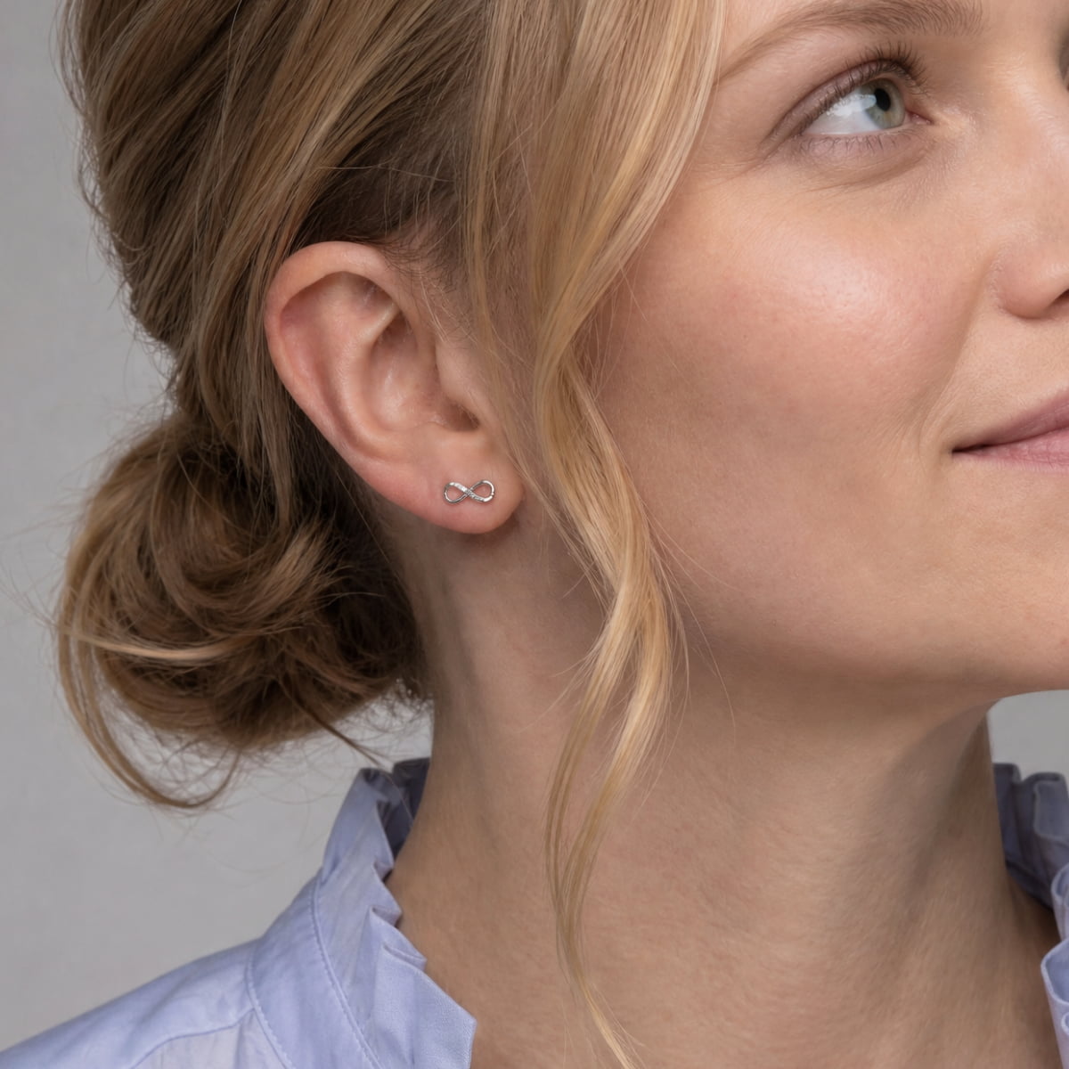 A close-up of a woman's ear wearing a tiny silver infinity symbol stud earring set with lab grown diamonds.