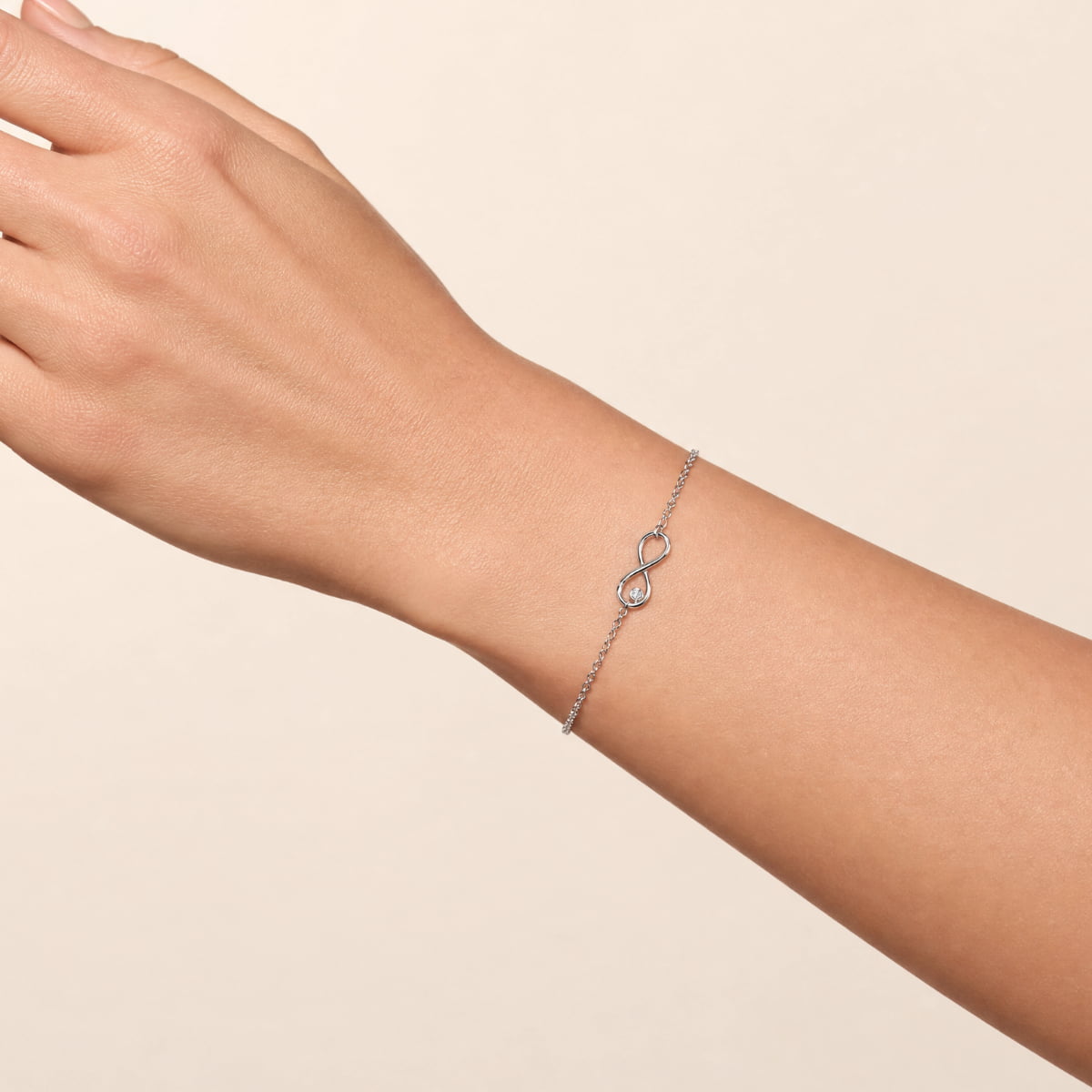 A delicate silver infinity bracelet with a small solitaire lab grown diamond, shown worn on a person's wrist against a warm beige background.