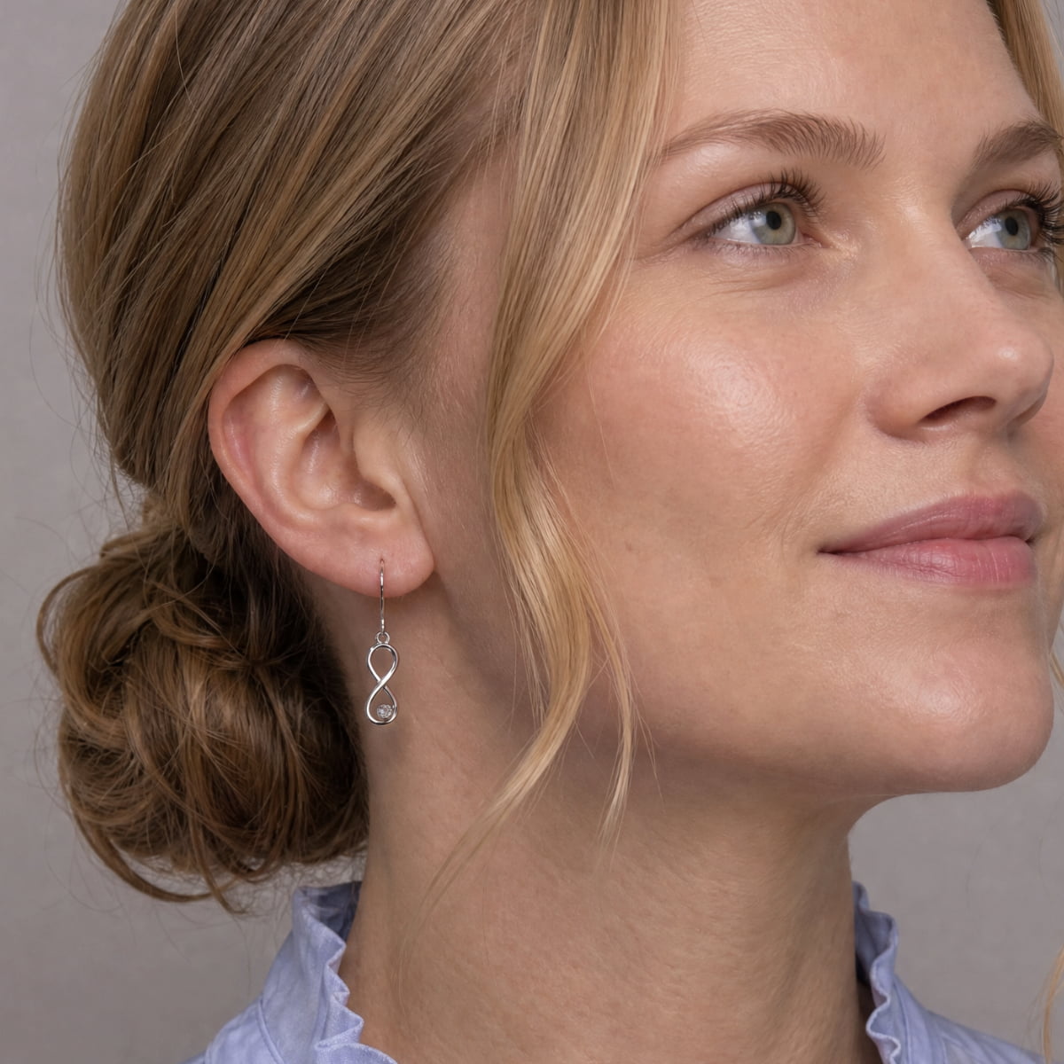 A close-up of a woman wearing a delicate silver infinity-shaped drop earring, set with a lab grown diamond.