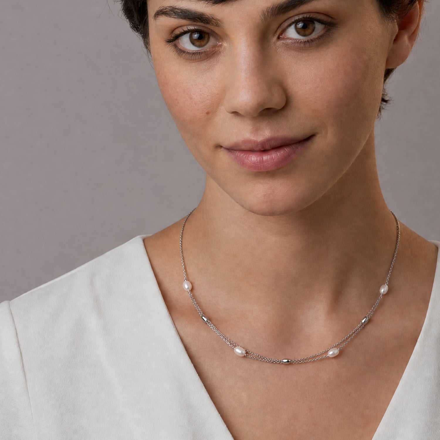 A close-up of a woman wearing a delicate silver chain necklace with small white pearls and silver beads spaced along the chain, against a plain gray background.