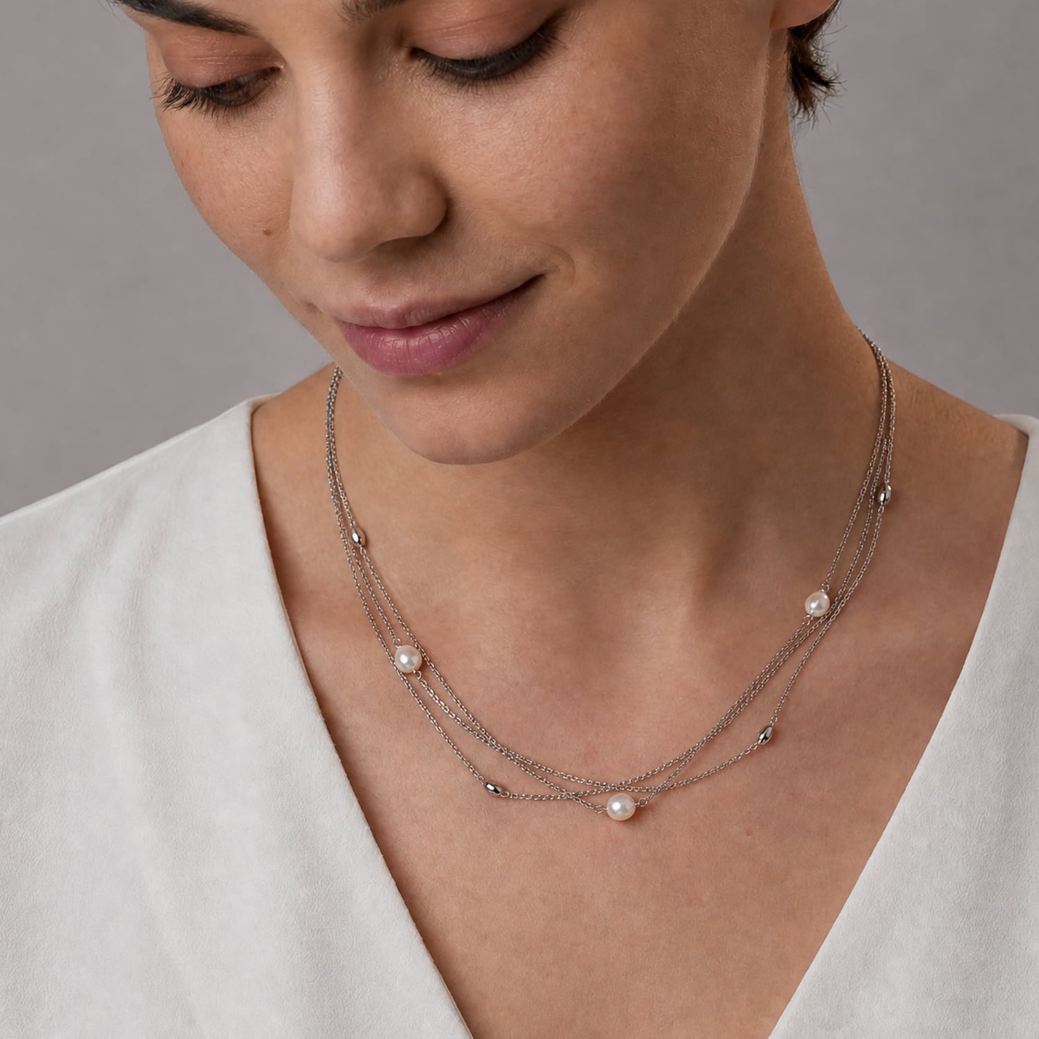A close-up of a woman wearing a delicate triple-layered silver necklace with small silver beads and white pearl accents, paired with a white V-neck top.
