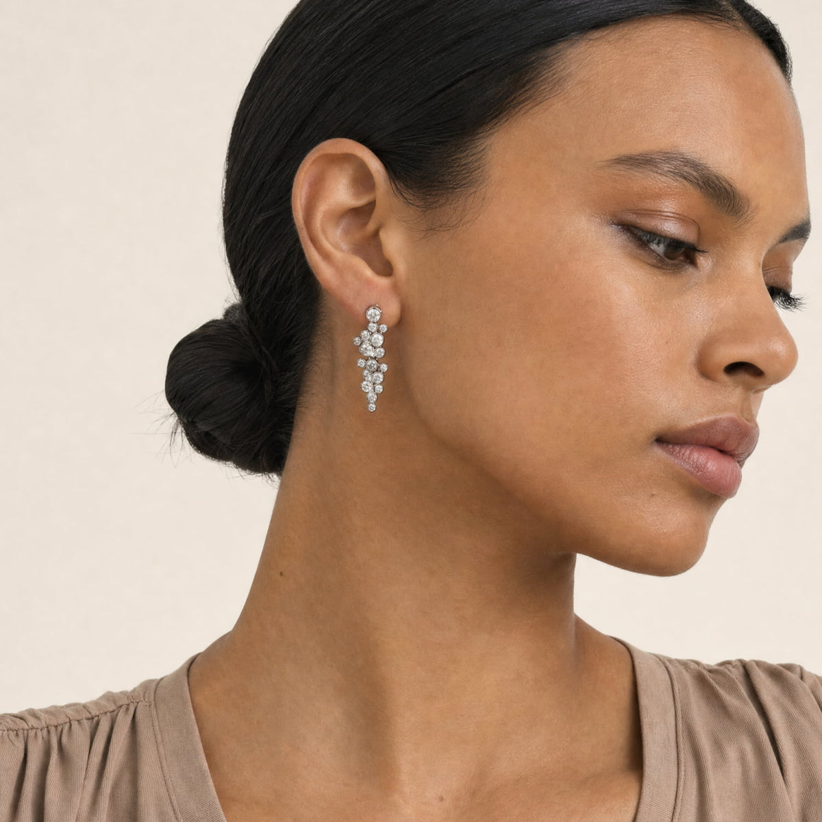 A close-up side profile of a woman with dark hair in a bun, wearing a silver, multi-stone drop earring with clear cubic zirconia, set against a plain beige background.