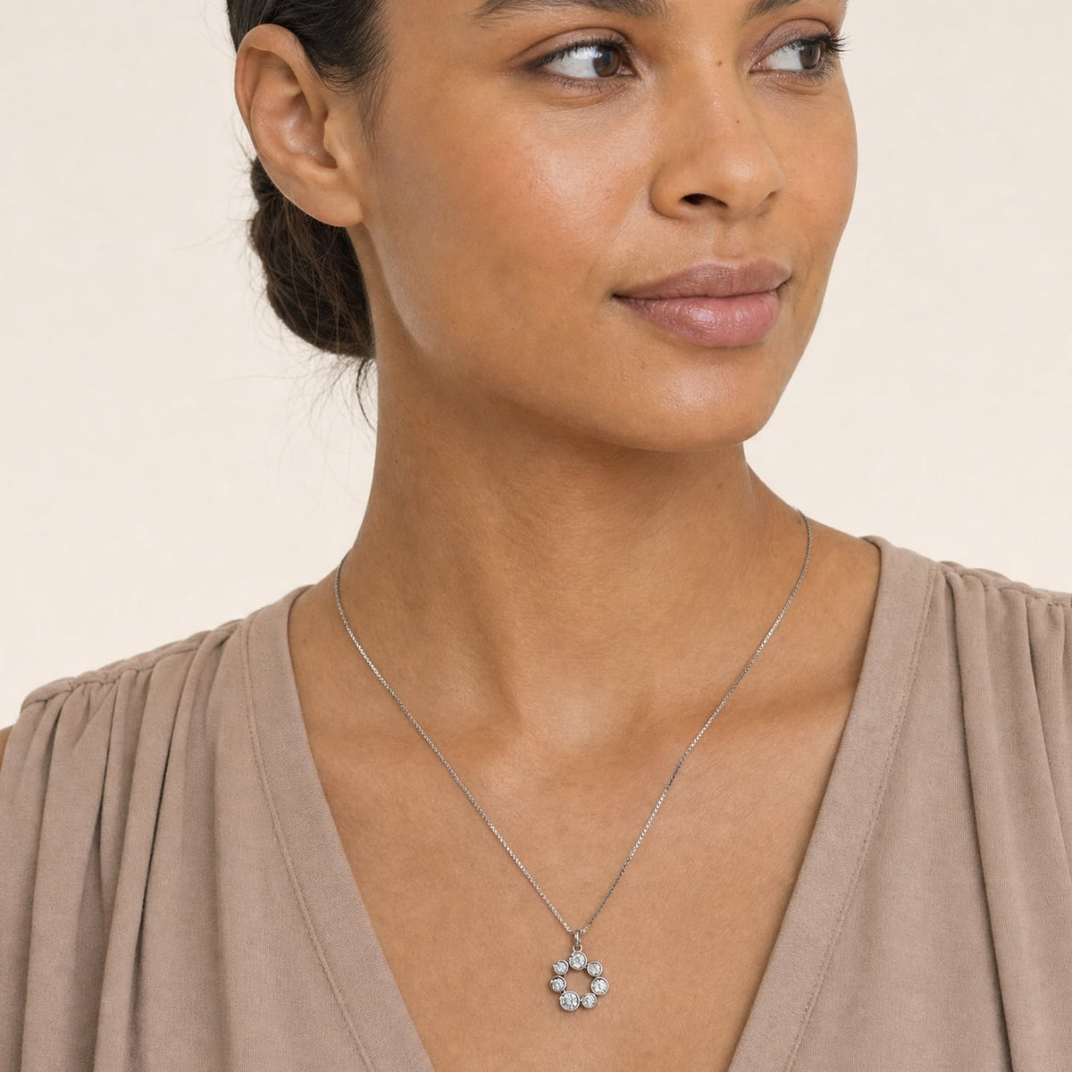A close-up of a woman wearing a silver chain necklace with a small flower-shaped pendant featuring clear stones, paired with a taupe V-neck top against a light beige background.