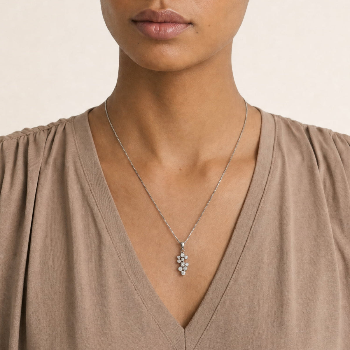 A close-up of a model wearing a silver necklace with a grape cluster pendant, styled with a taupe V-neck top against a light beige background.