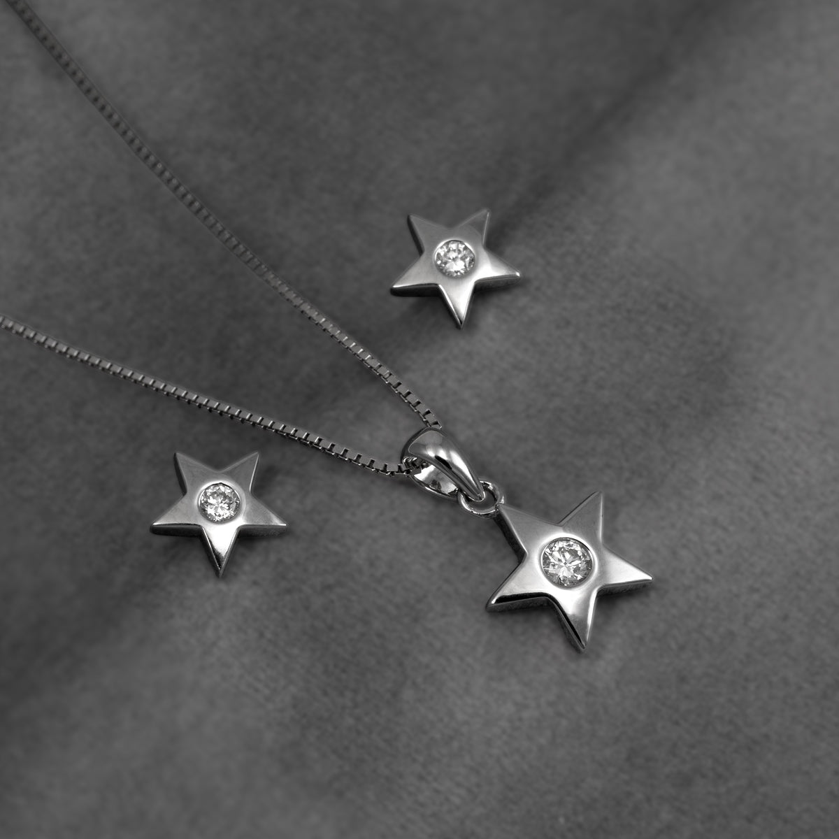 Silver star pendant and matching stud earring, all with clear lab grown diamonds, displayed on a dark gray textured fabric background.