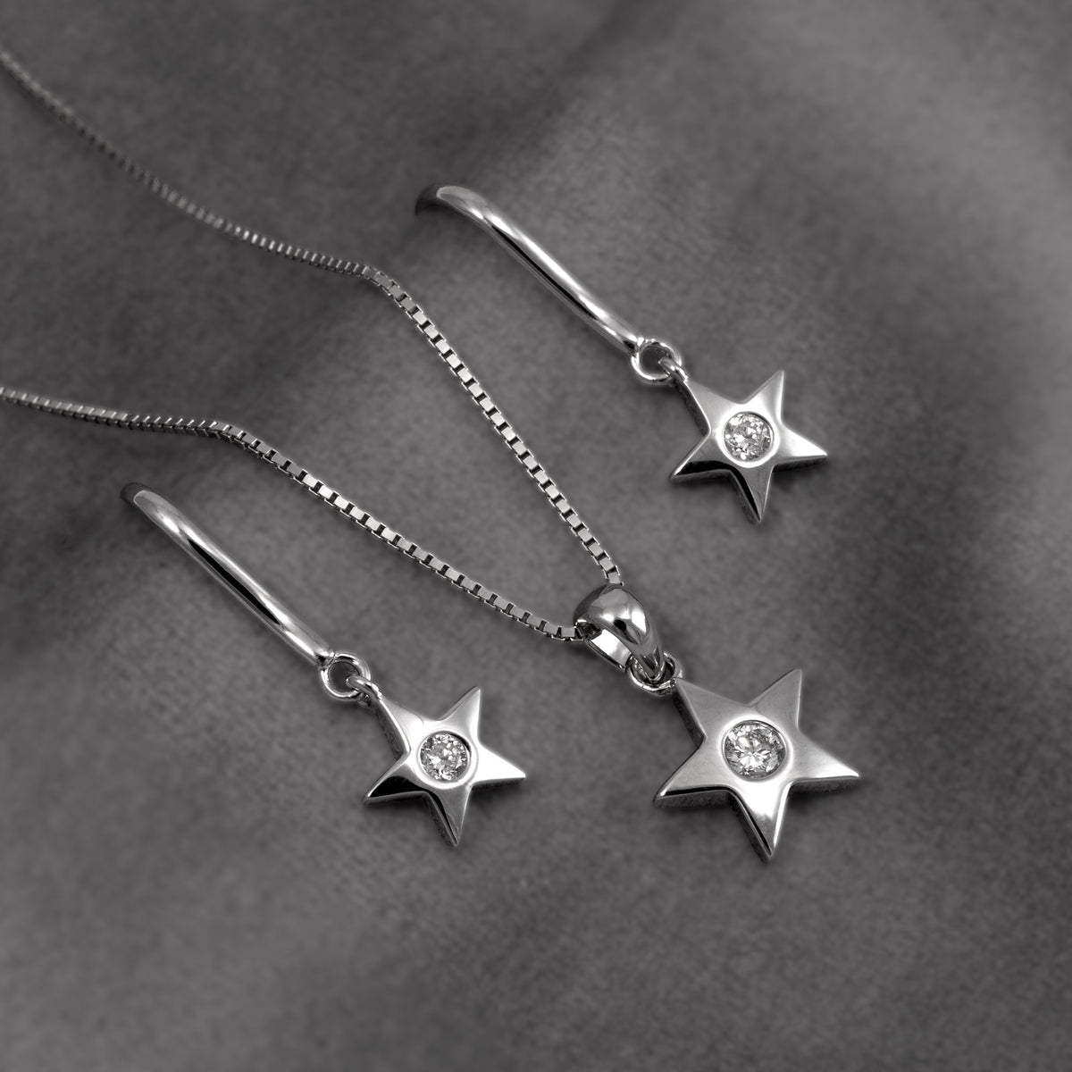 Silver star pendant and matching drop earring, all with clear lab grown diamonds, displayed on a dark gray textured fabric background.