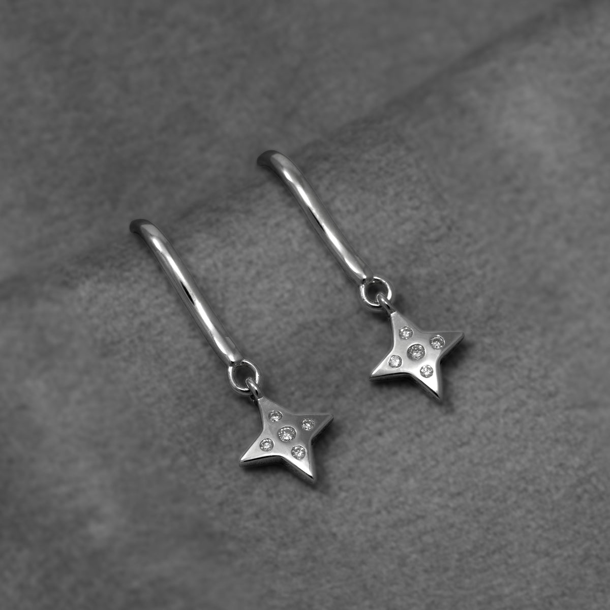 A pair of silver drop earrings featuring a four-pointed star charm with lab grown diamonds, suspended from curved hook ear wires, shown on a grey background.