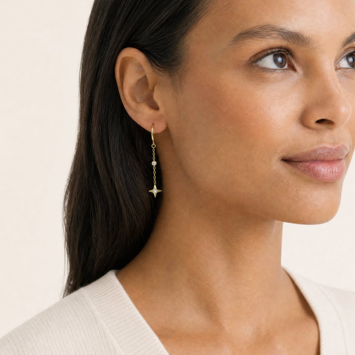 A close-up of a woman wearing a delicate gold vermeil drop earring with a small star charm and lab diamond accents, set against a soft beige background.