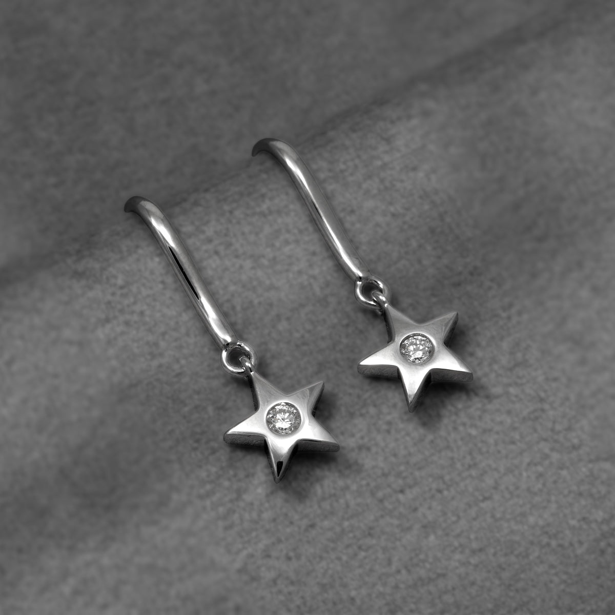 A pair of silver star-shaped drop earrings with a lab grown diamond in the center of each star, shown on a textured gray fabric background.