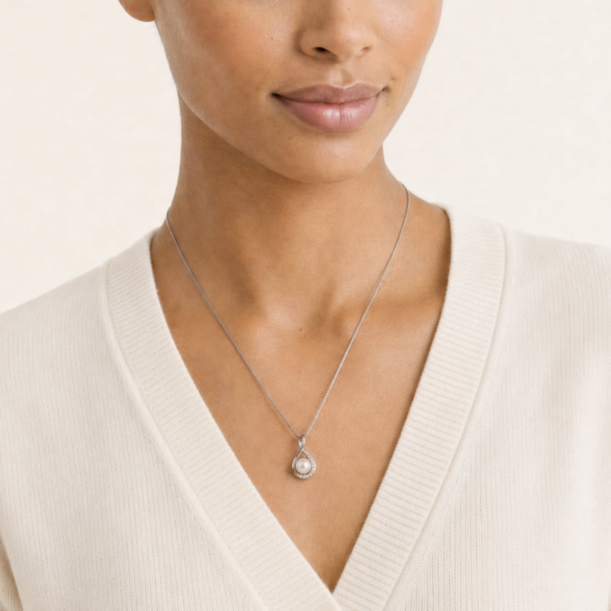 A close-up of a model wearing a silver infinity-shaped pendant with a freshwater pearl and lab grown diamonds, paired with a cream-colored ribbed V-neck cardigan.
