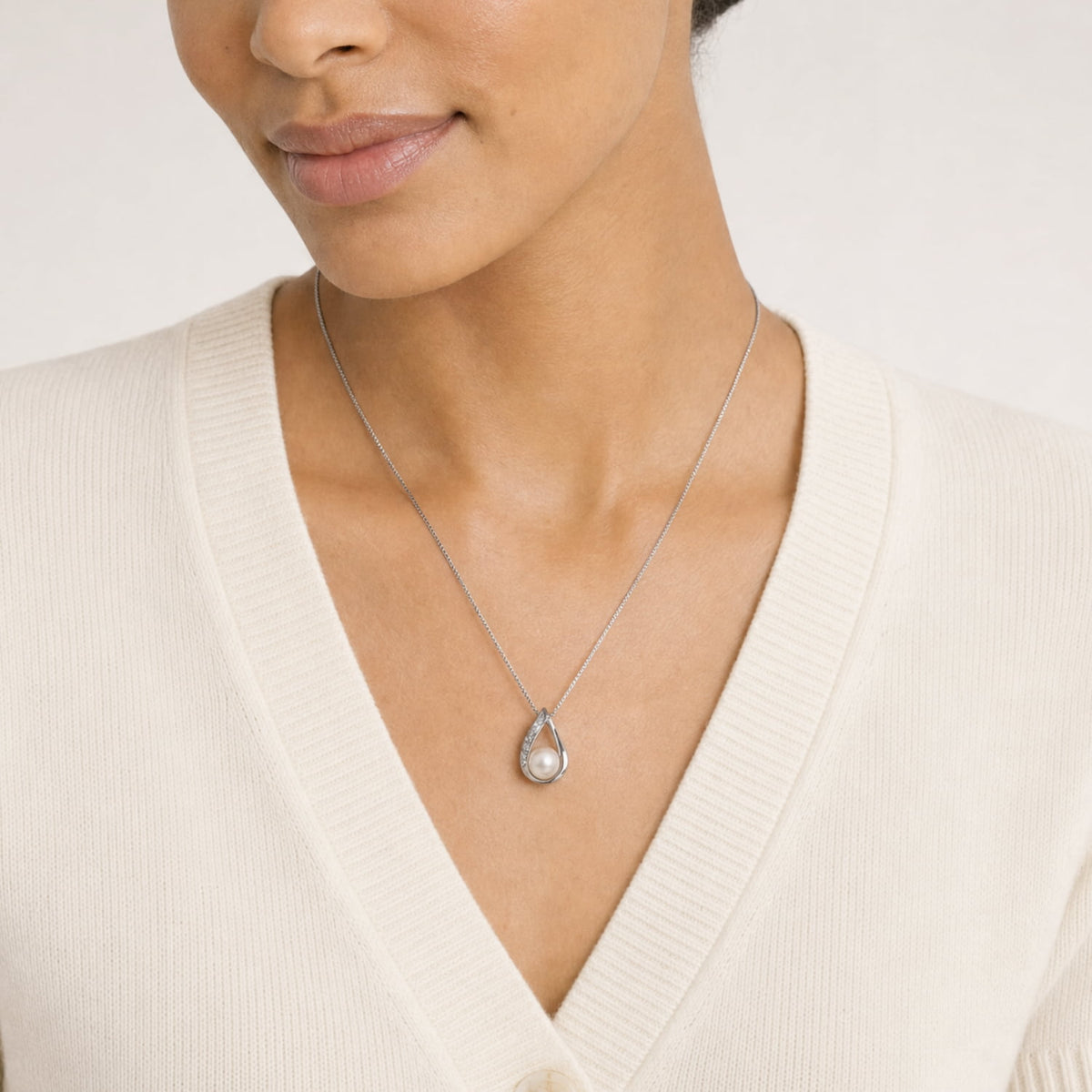 A close-up of a model wearing a delicate silver teardrop pendant necklace with a freshwater pearl, paired with an off-white ribbed V-neck sweater.