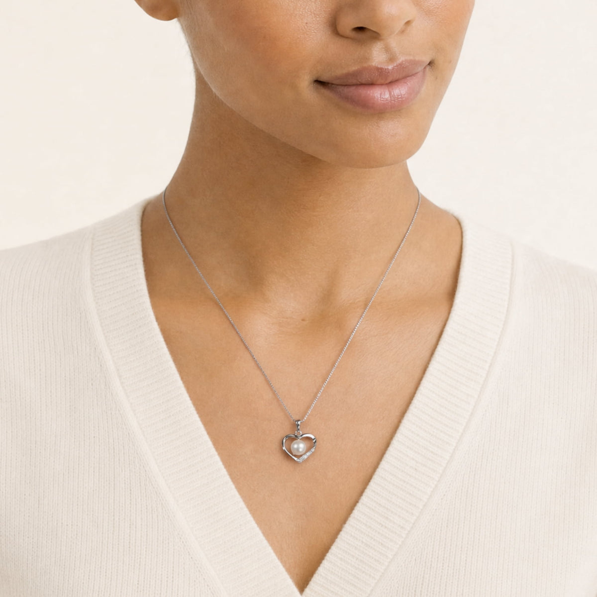 A close-up of a model wearing a delicate silver heart-shaped pendant with a freshwater pearl and lab grown diamonds, styled with a cream-colored V-neck sweater.