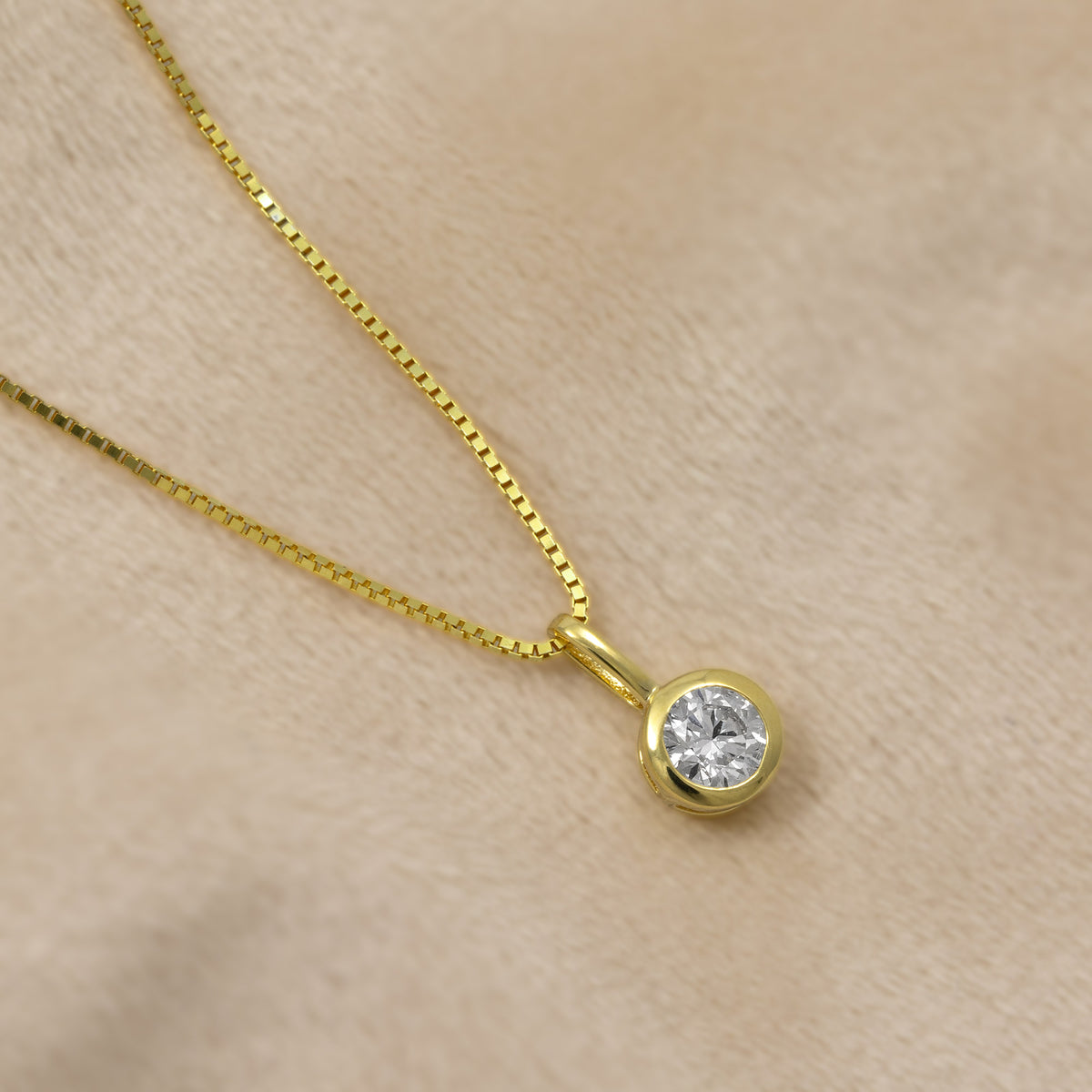 A gold box chain necklace with a round bezel-set half carat diamond pendant, displayed on a soft beige textured background.