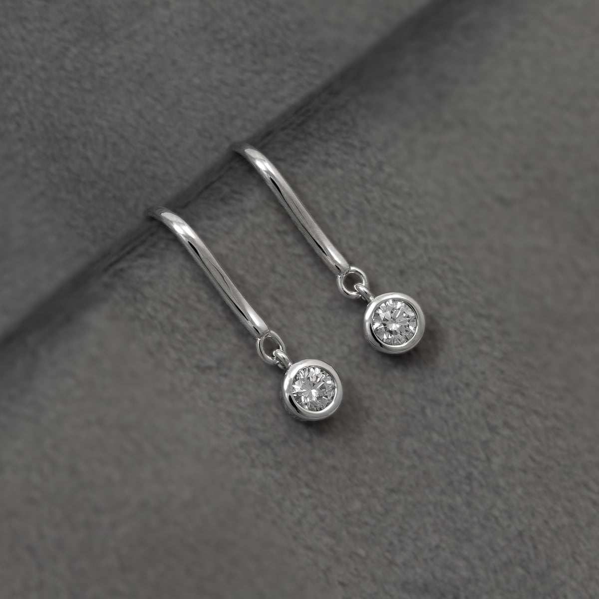 A pair of silver hook earrings, each featuring a quarter carat round bezel-set lab diamond, shown against a grey background.