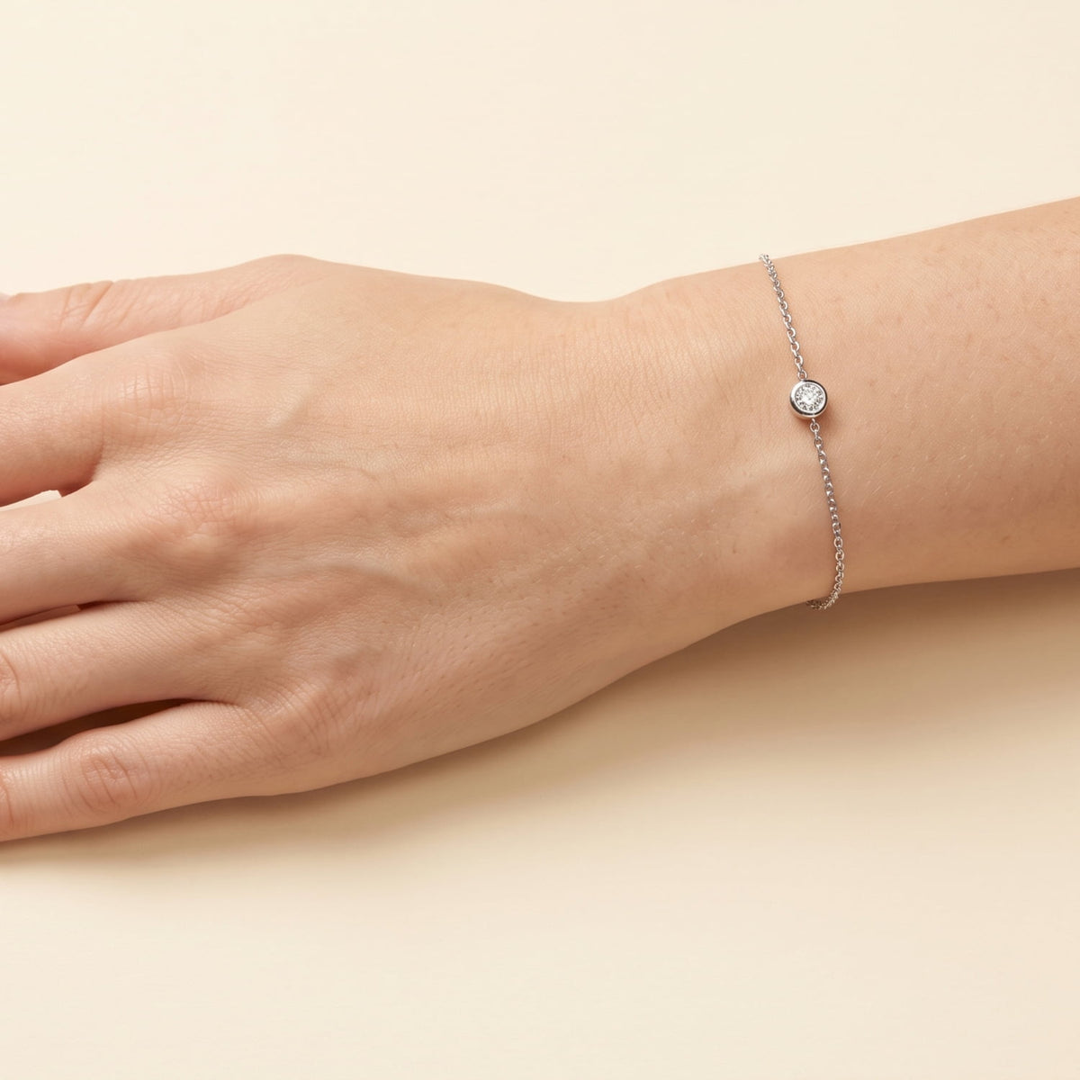A delicate silver chain bracelet with a bezel-set lab diamond, shown worn on a person's wrist against a warm beige background.